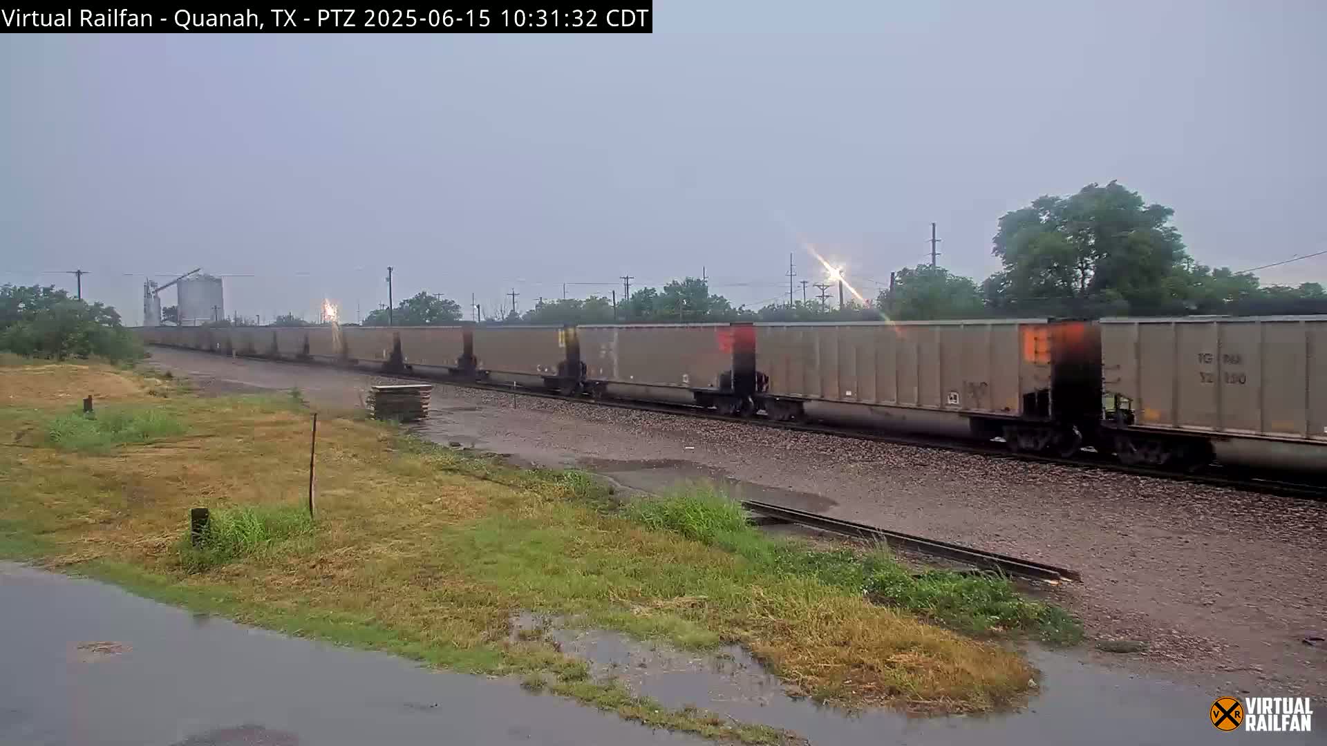 A long freight train moves along a track in overcast conditions, with puddles visible on the ground.