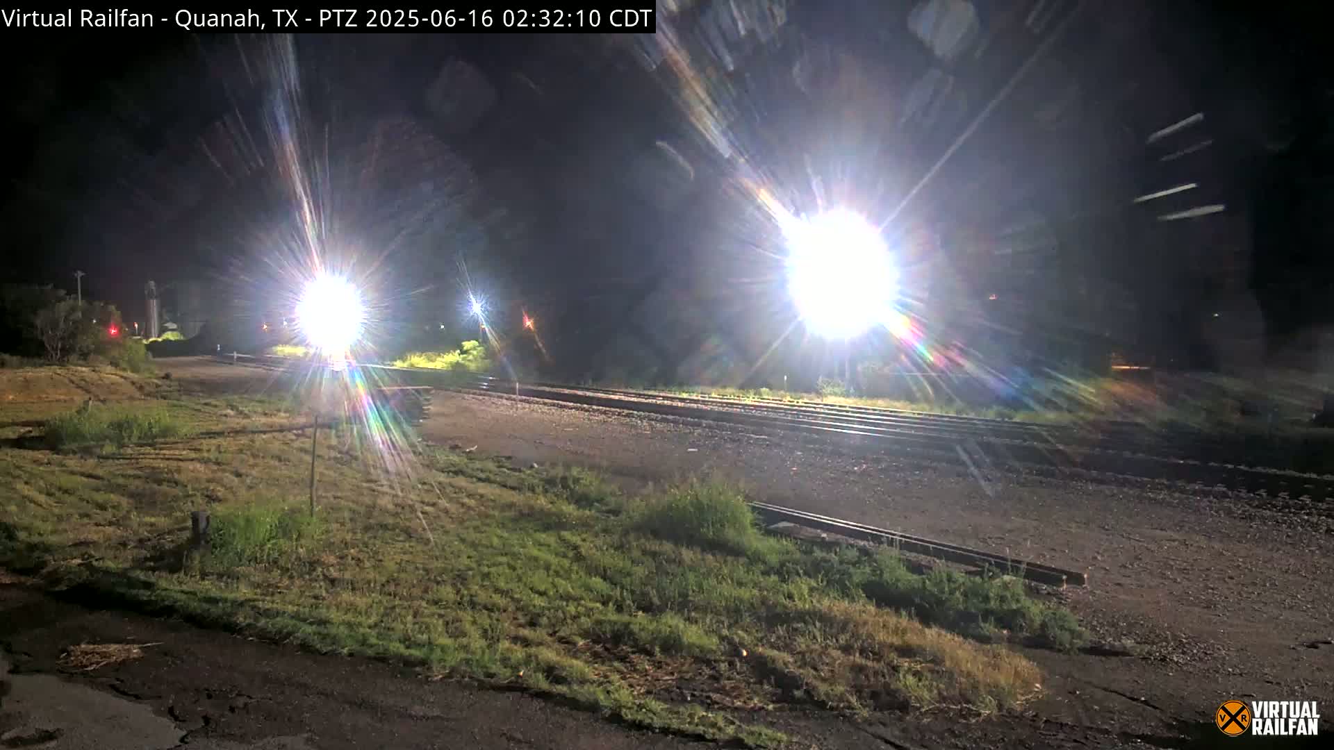 At night, bright lights illuminate a section of railroad tracks bordered by grassy areas.
