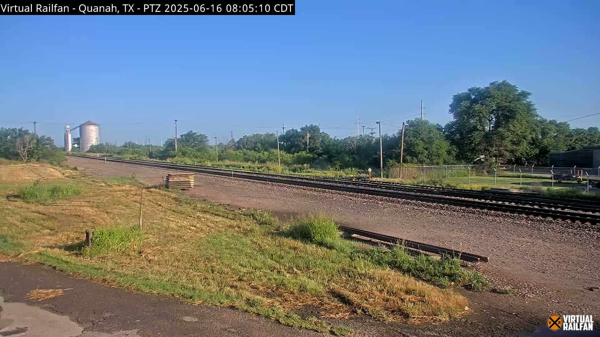 A train track runs through a grassy area next to a grain silo under a clear sky.