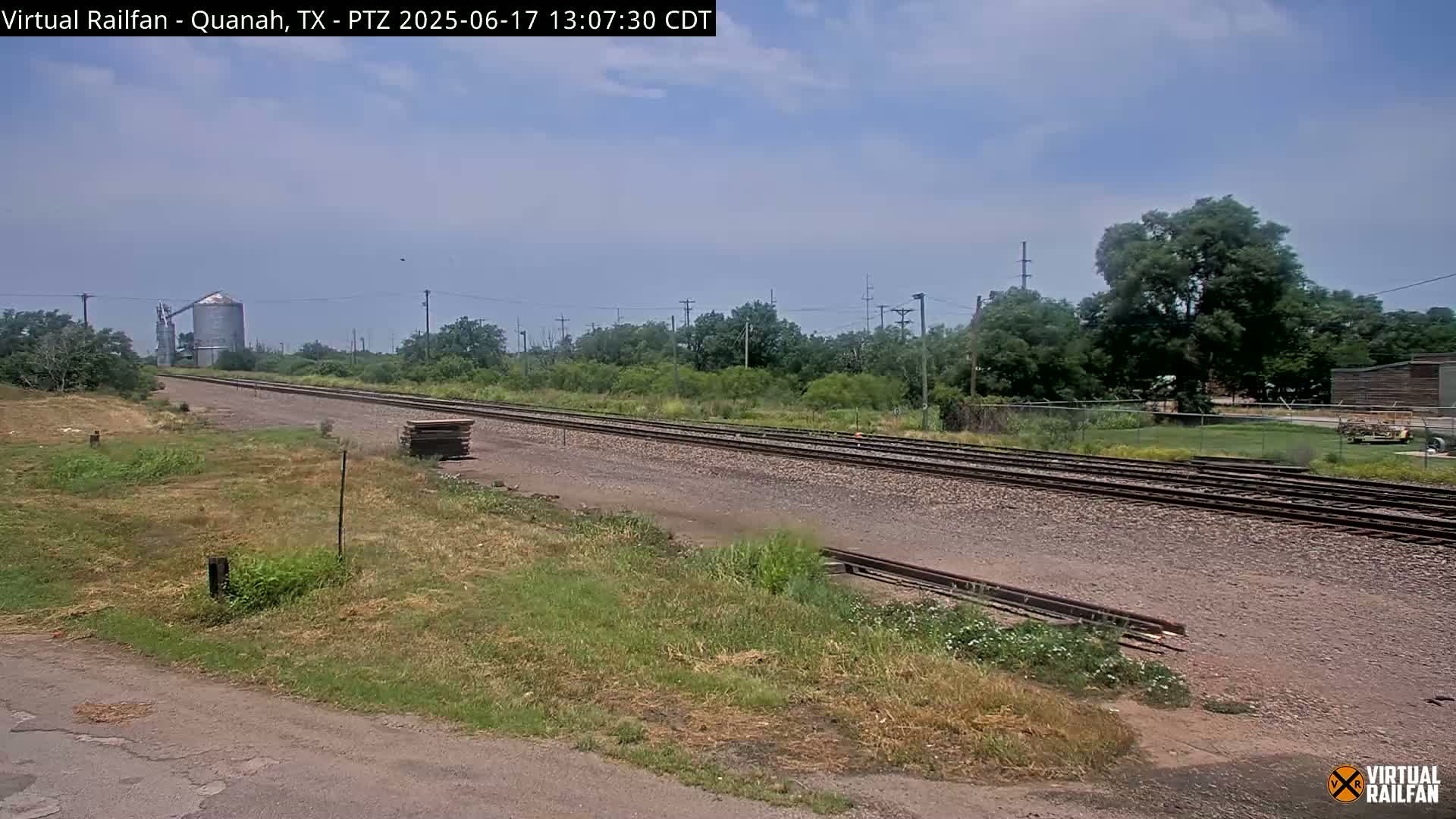 A view of a railway track stretching into the distance under a partly cloudy sky, with dry, grassy land on either side, a grain silo in the background, and some trees and buildings visible in the far distance.