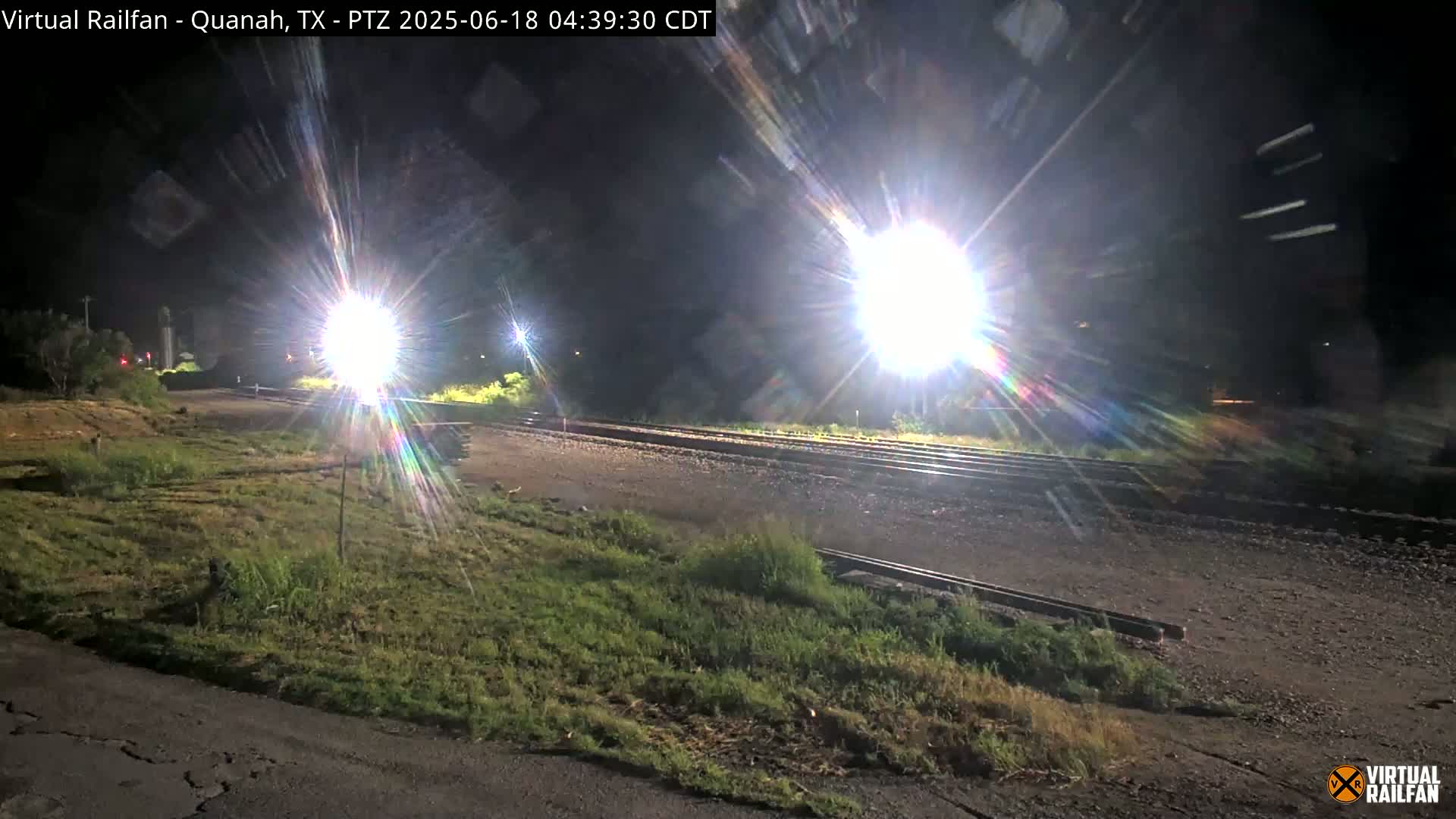 A nighttime view of train tracks illuminated by bright lights, with grassy areas visible on either side.