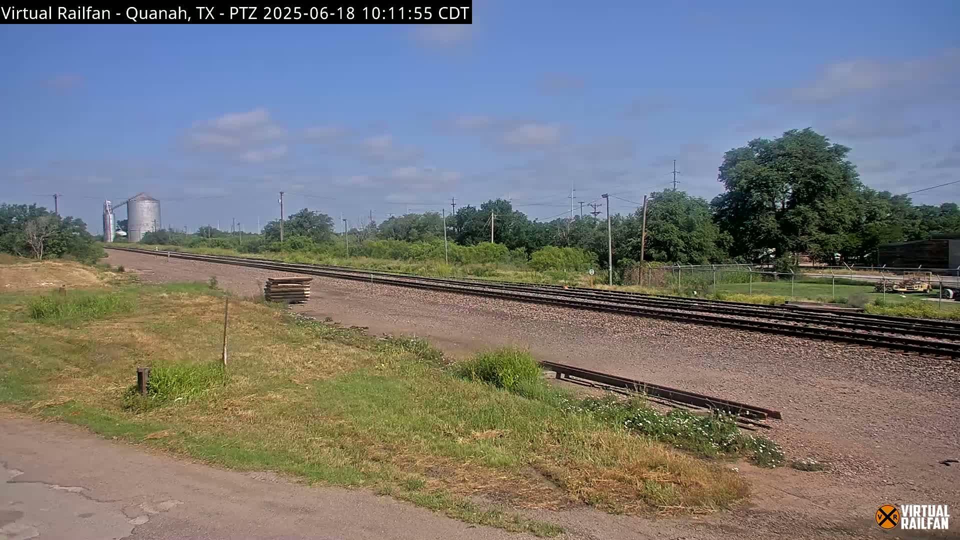 A long stretch of railroad tracks extends into the distance under a partly cloudy sky, passing alongside grassy fields and a grain silo.