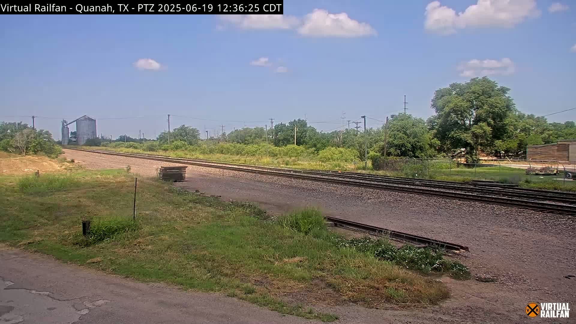 A mostly sunny day shows a view of railroad tracks stretching into the distance, bordered by grassy fields and trees, with a grain silo visible in the background.