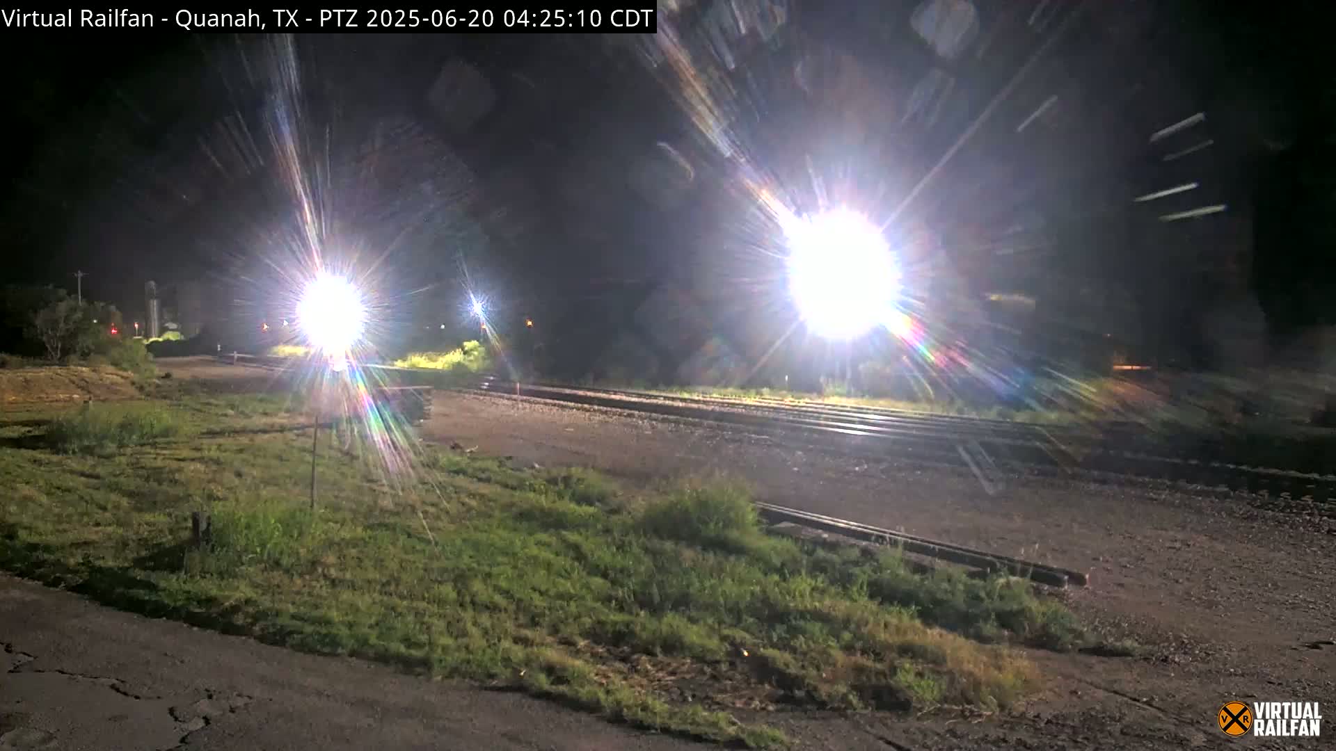 At night, bright lights illuminate a section of railroad tracks next to a grassy area.
