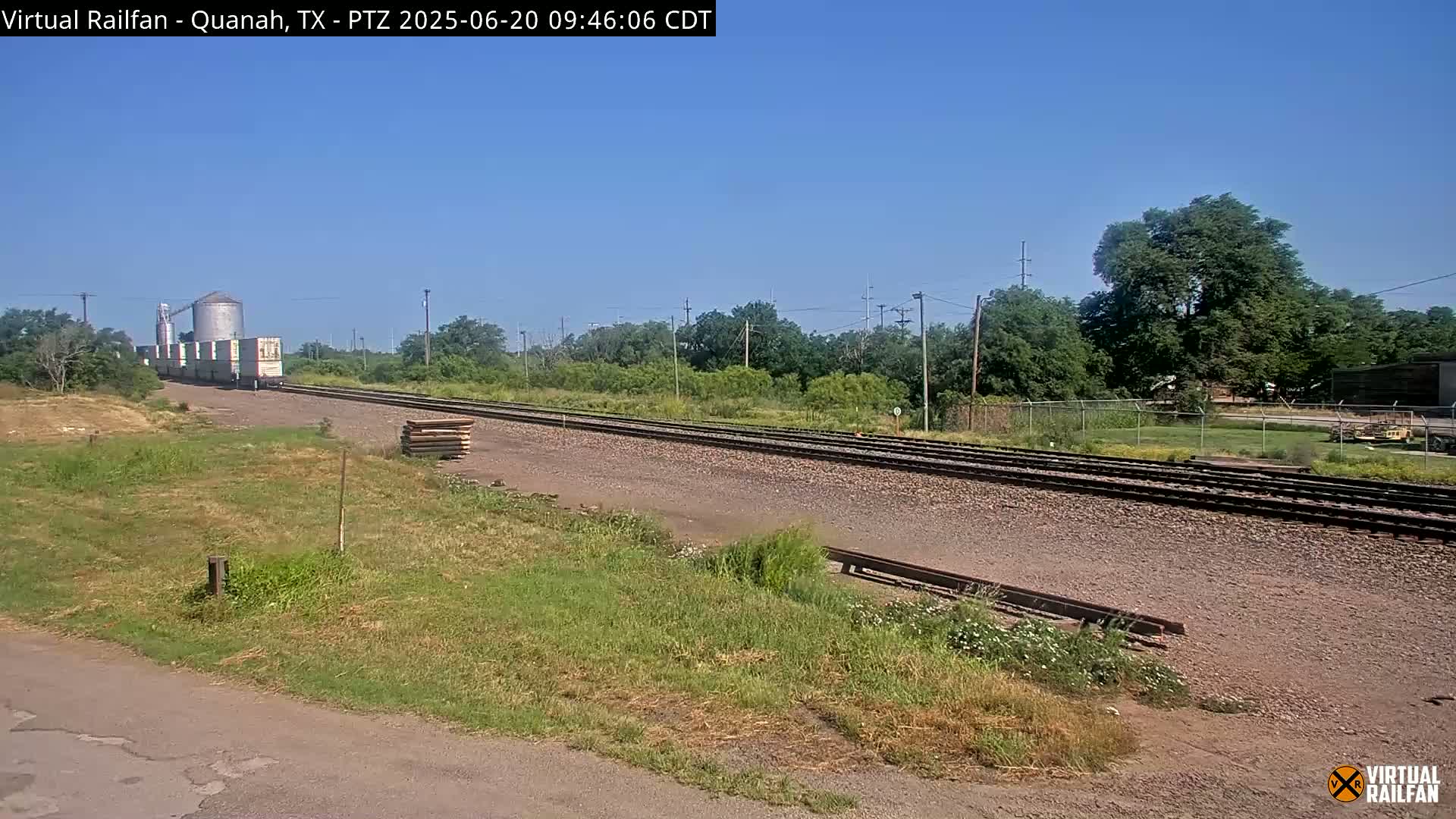 A train carrying several boxcars passes by a grain silo and railroad tracks on a clear, sunny day.