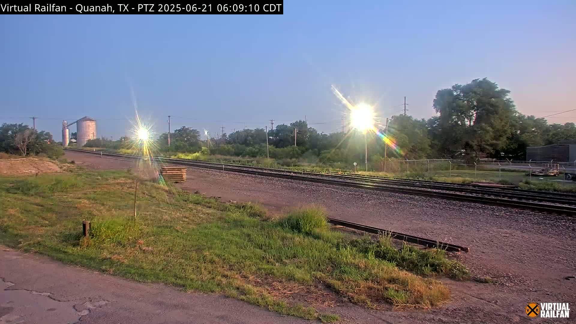 A sunrise illuminates a railway track that stretches toward a grain silo and distant trees under a clear sky.