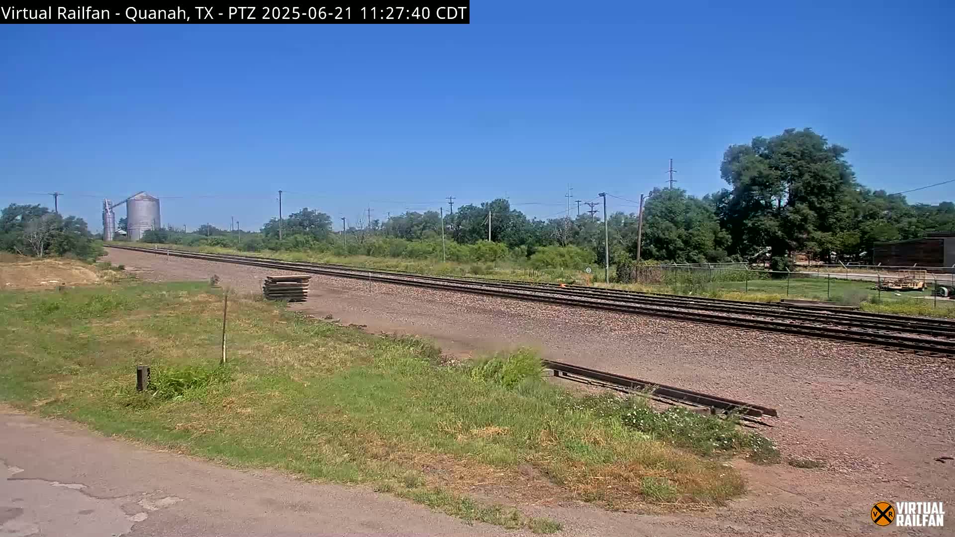A long stretch of railroad tracks runs through a grassy area under a clear blue sky, with trees and a grain silo visible in the distance.