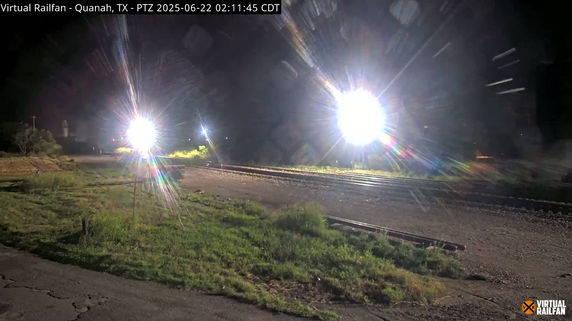 At night, brightly lit train tracks are visible, bordered by patches of grass and a dark roadway.