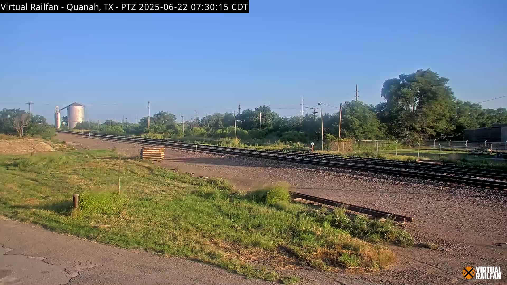 A clear, sunny day reveals a view of train tracks extending into the distance, passing by a grassy area with a distant silo and some trees.