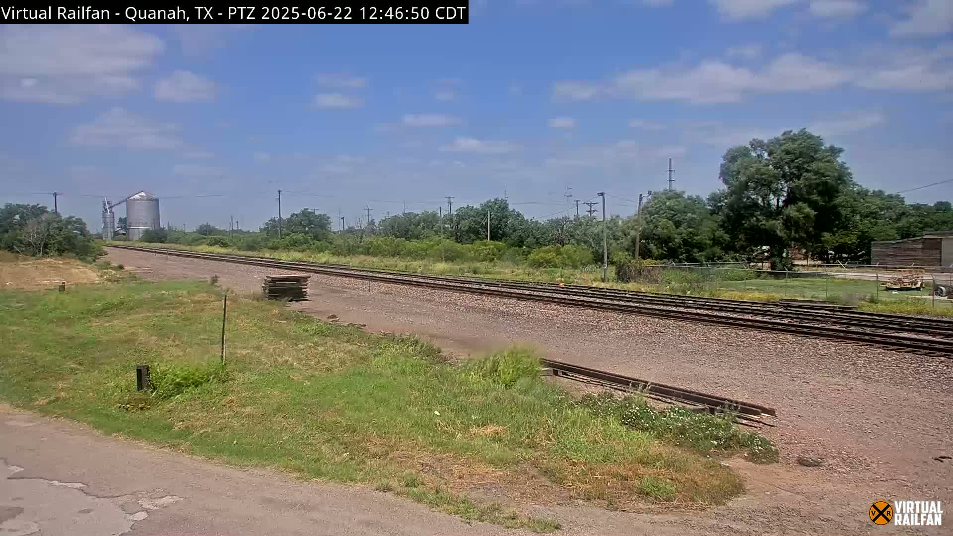 A long stretch of railroad tracks extends into the distance under a partly cloudy blue sky, passing alongside grassy fields and a grain silo.