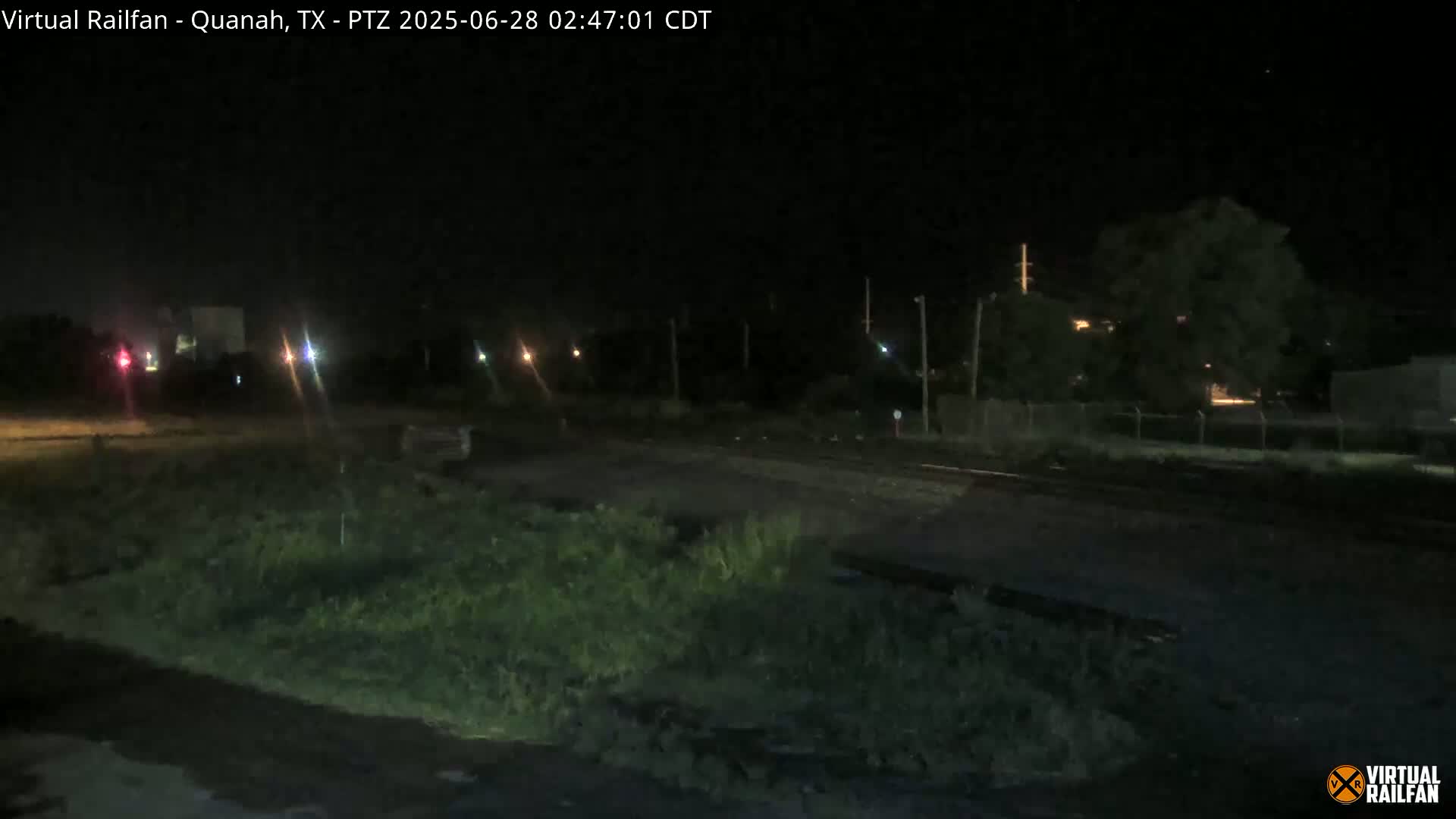 A nighttime view of a railroad crossing with overgrown vegetation in the foreground, under a clear, dark sky.