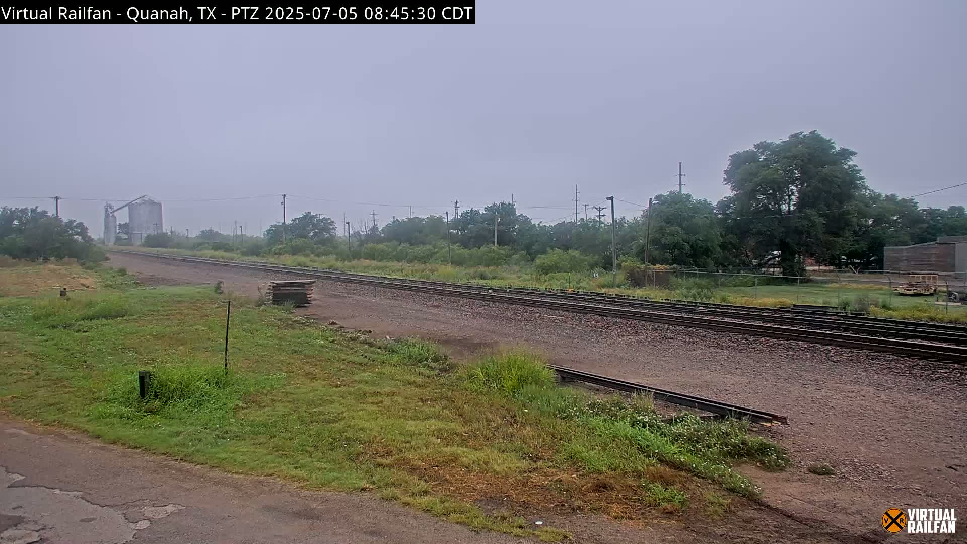 A mostly clear, hazy day shows a view of railroad tracks extending into the distance past a grassy area with trees and a grain silo in the background.
