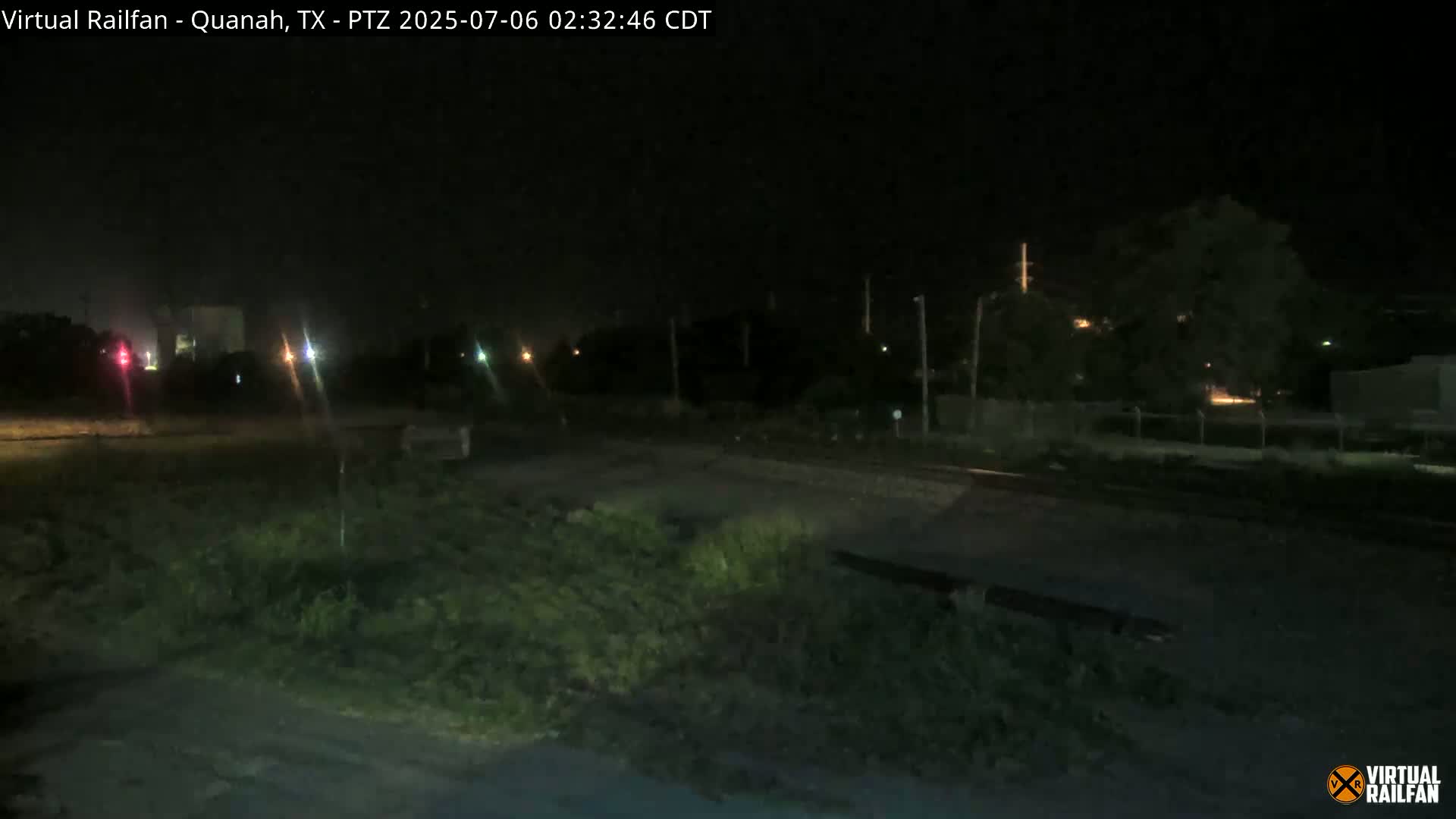 A nighttime view of a mostly dark, grassy area next to railroad tracks, with a few distant lights visible.
