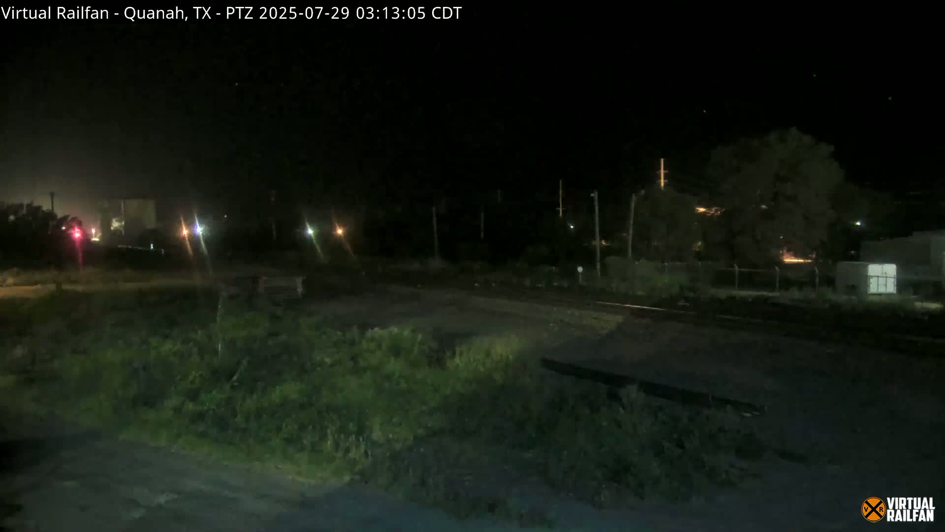 A nighttime, low-light view of a railroad crossing, with vegetation in the foreground and distant lights visible.