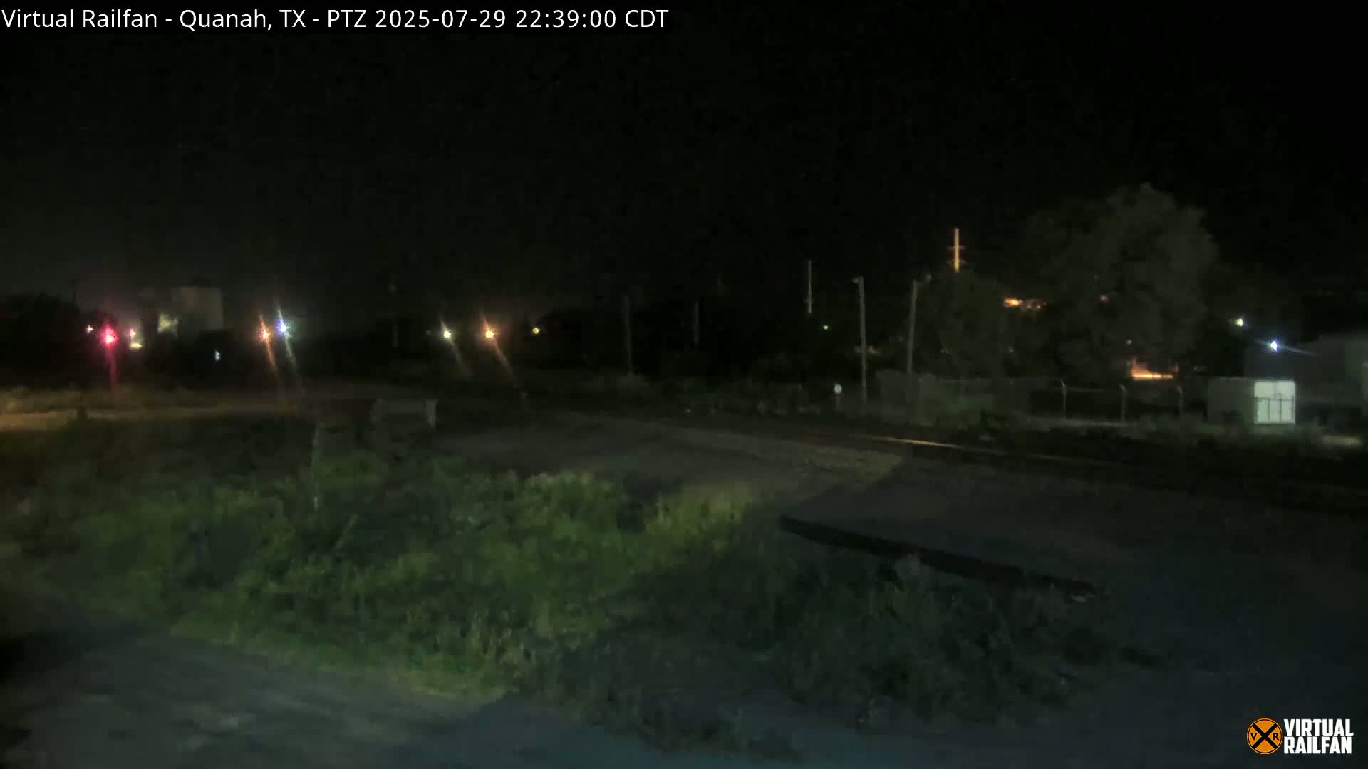 A nighttime, low-light view of a railroad crossing, with vegetation in the foreground and distant lights visible.