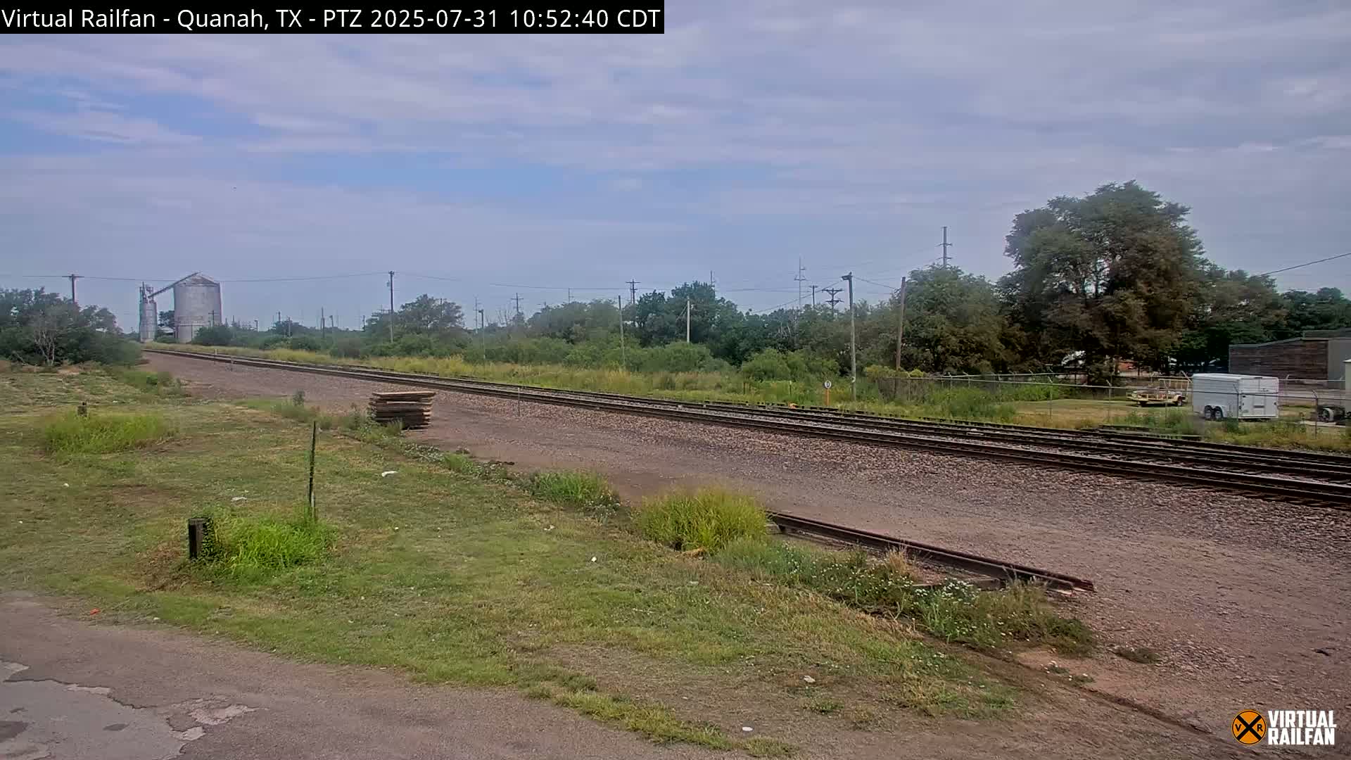 A long stretch of train tracks extends into the distance under a partly cloudy sky, passing beside a grassy area with some vegetation and a grain silo visible in the far background.