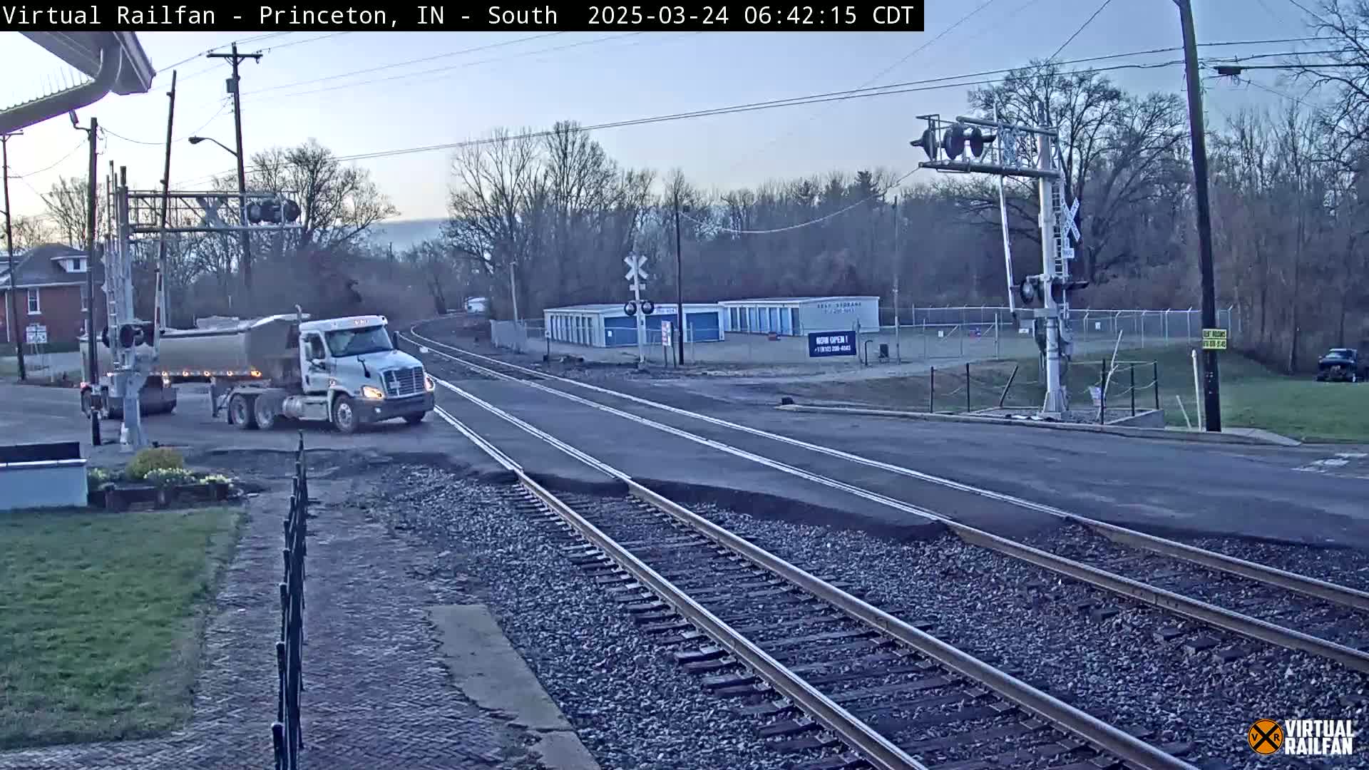 A white semi-truck with a tanker trailer is crossing a railroad intersection on a road under a twilight sky.