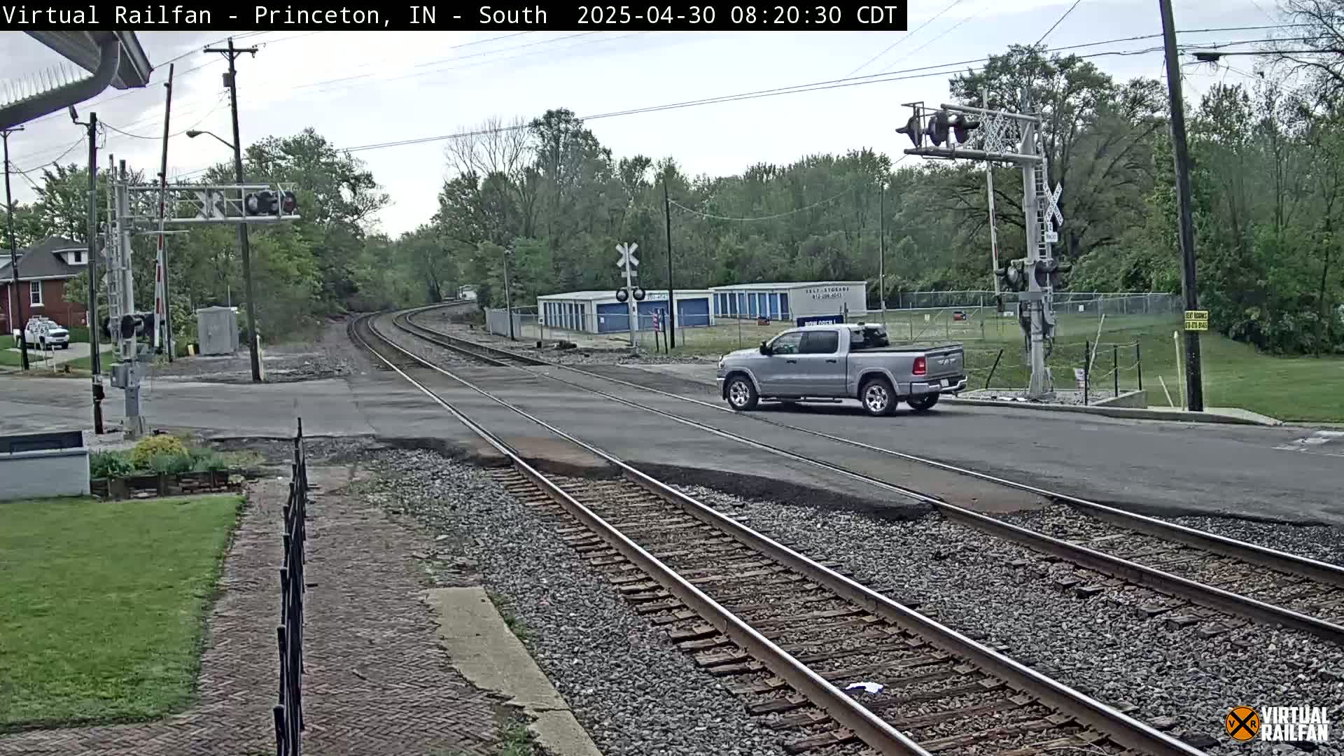 A silver pickup truck crosses a road intersecting with railroad tracks under an overcast sky.