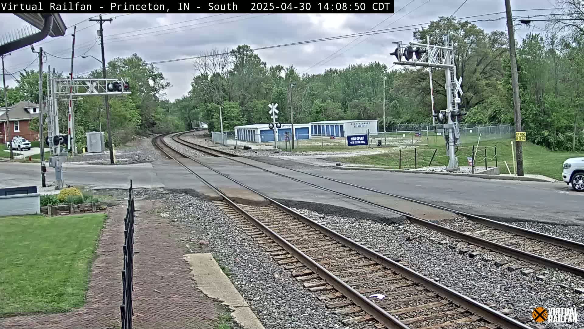 A railroad crossing intersects a road under an overcast sky, with trees and storage units visible in the background.