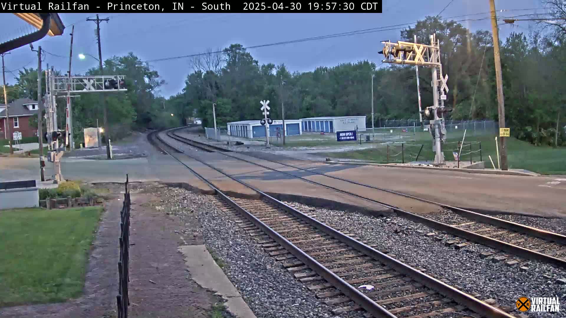 A railroad crossing at dusk shows two sets of train tracks intersecting a road, with a signal light and trees in the background.