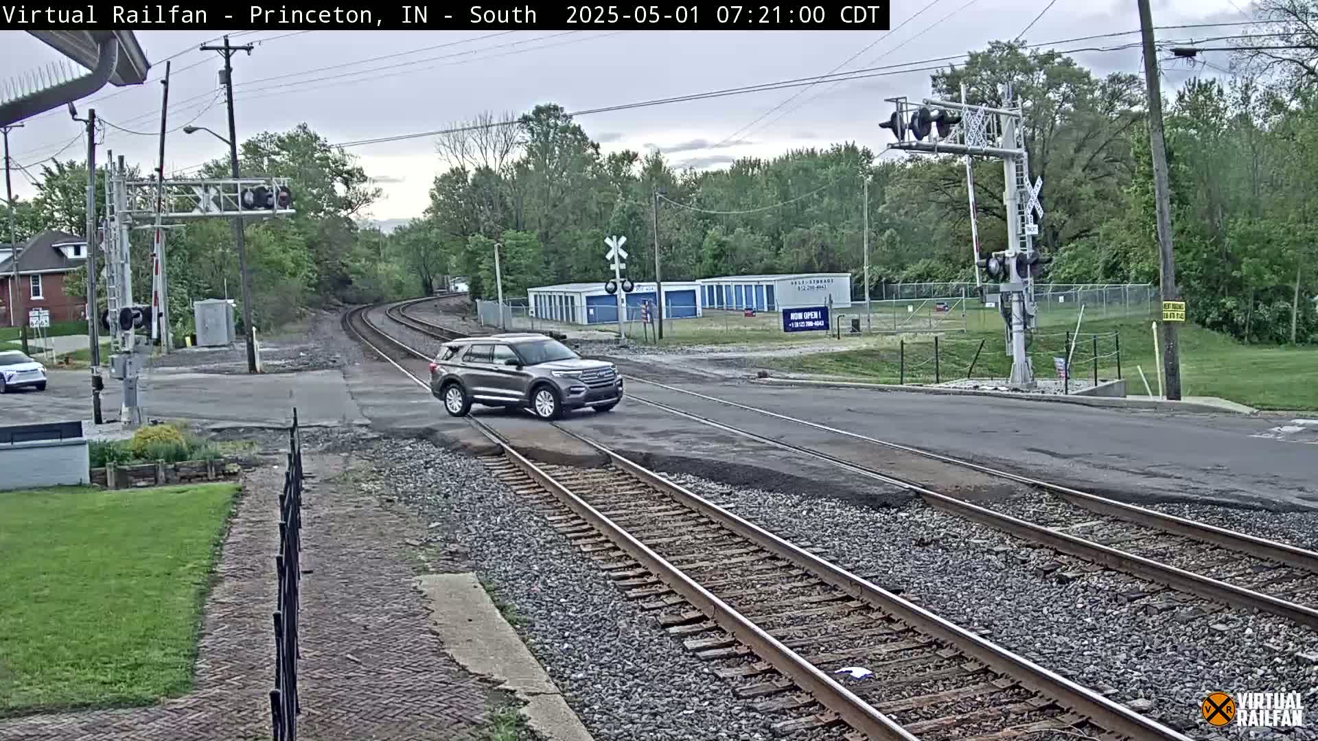 A gray SUV drives across a railroad crossing under an overcast sky, passing through a road intersection with railroad signals and trees in the background.