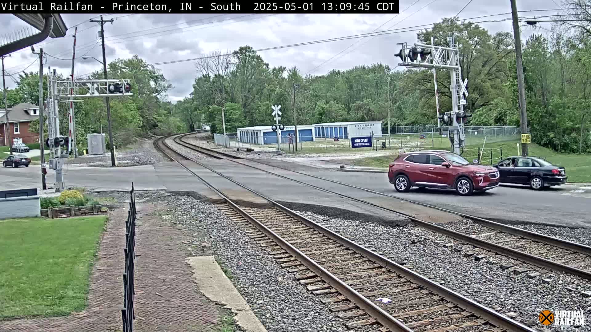 A railroad crossing with a maroon SUV and a black sedan stopped at the intersection under a partly cloudy sky.