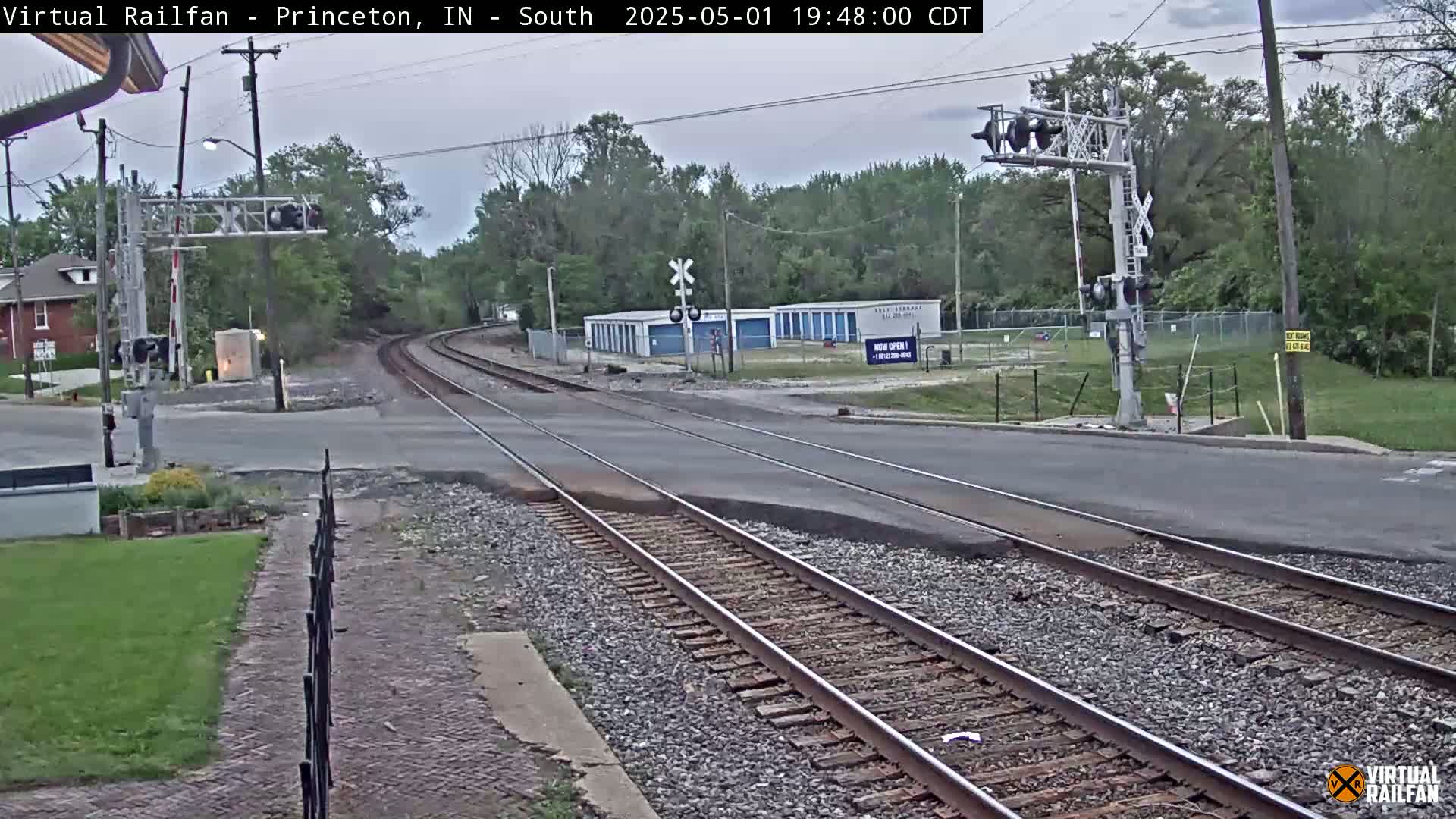 A railroad crossing intersects a street under an overcast sky, with trees and storage units visible in the background.