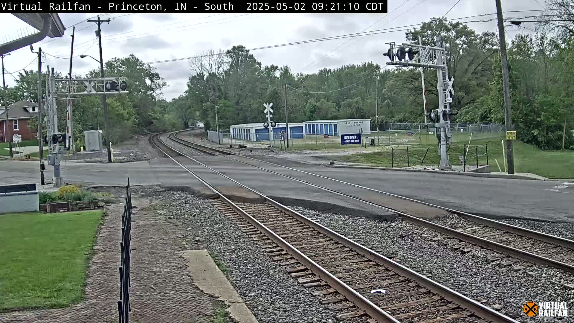 A railroad crossing in a residential area on an overcast day shows train tracks intersecting a road with signals and some storage units visible in the background.