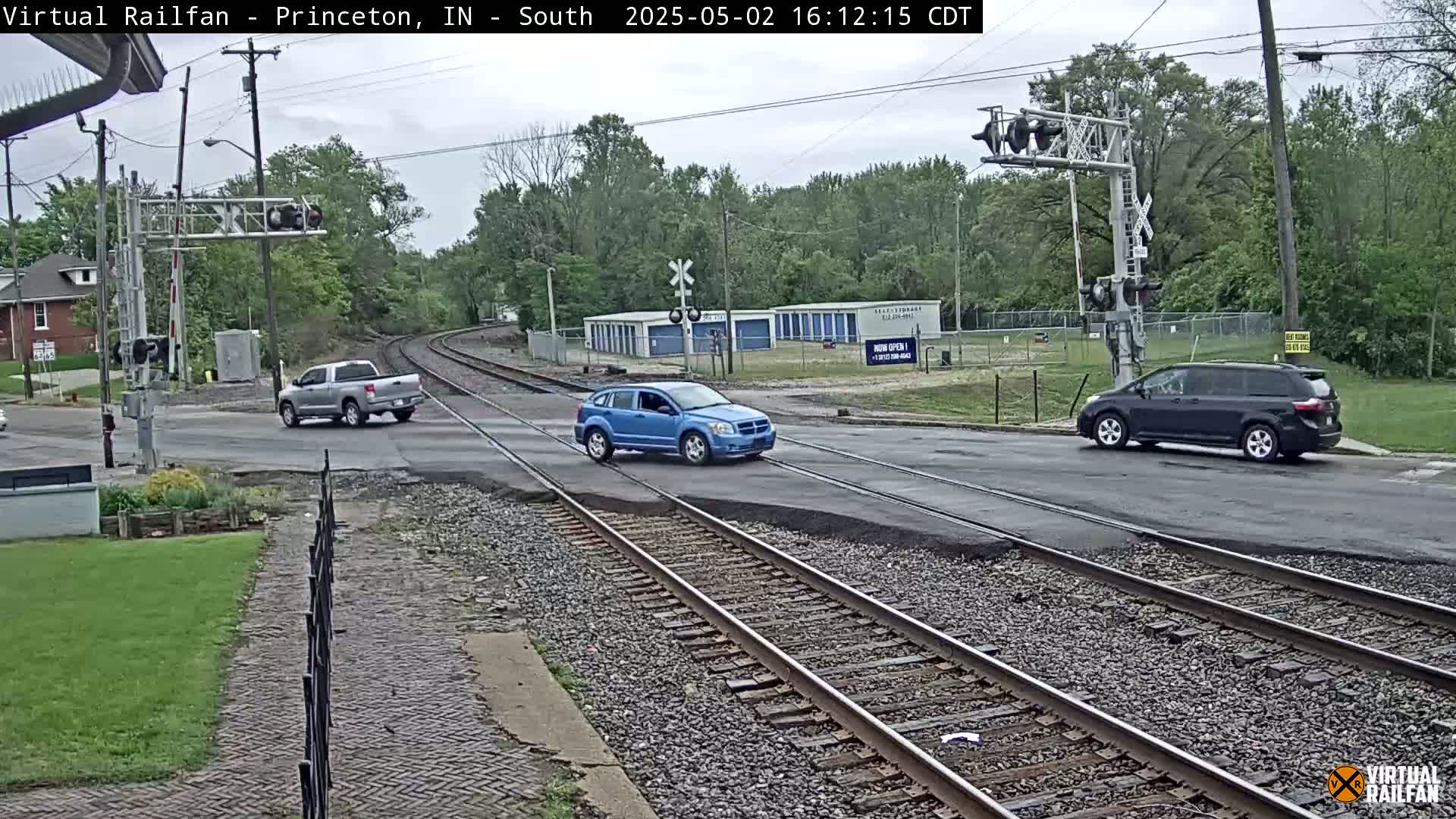 A street intersects railroad tracks under an overcast sky, with several vehicles crossing the intersection and trees lining the background.