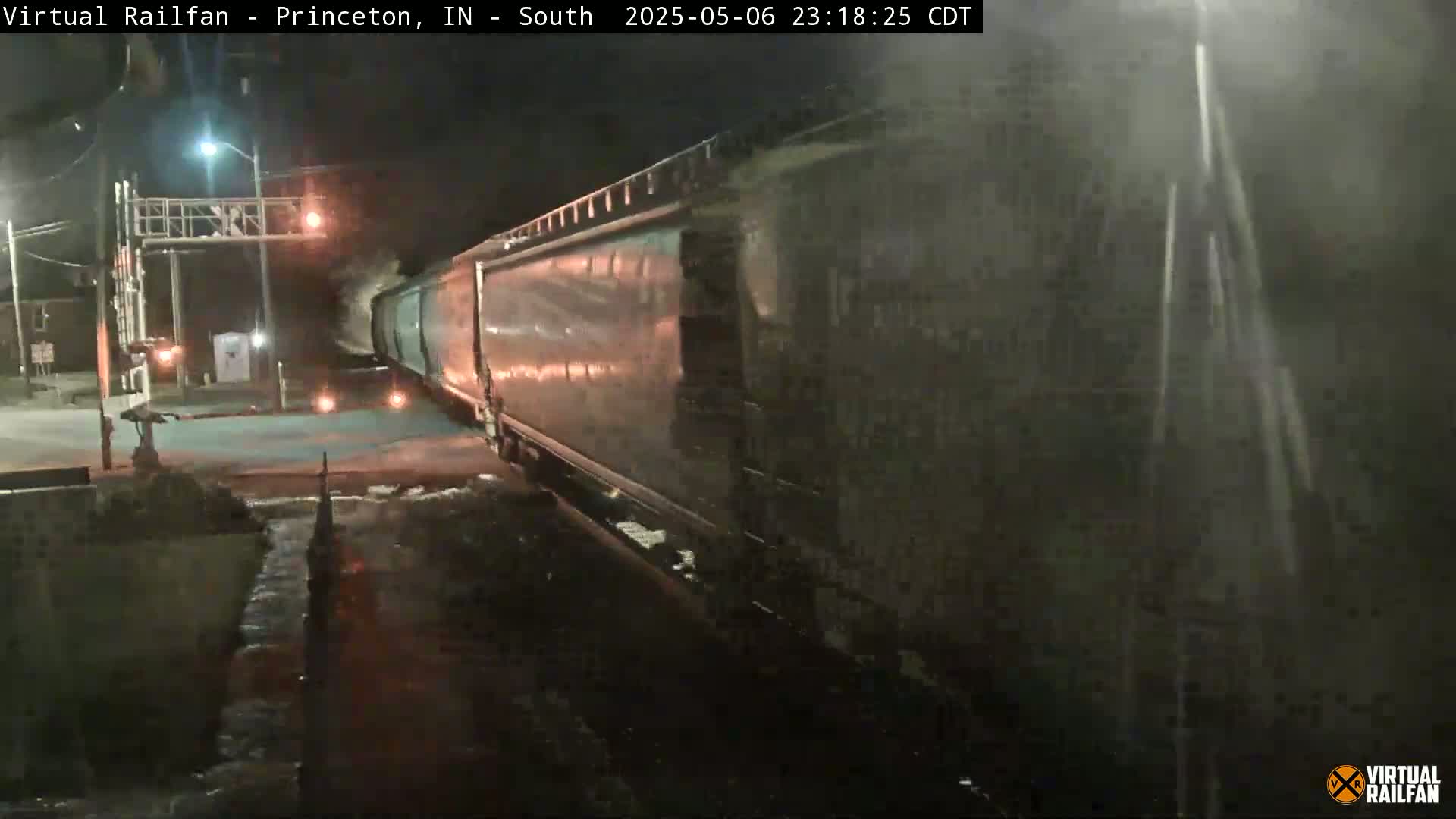 A long freight train passes through a nighttime railroad crossing in the rain.