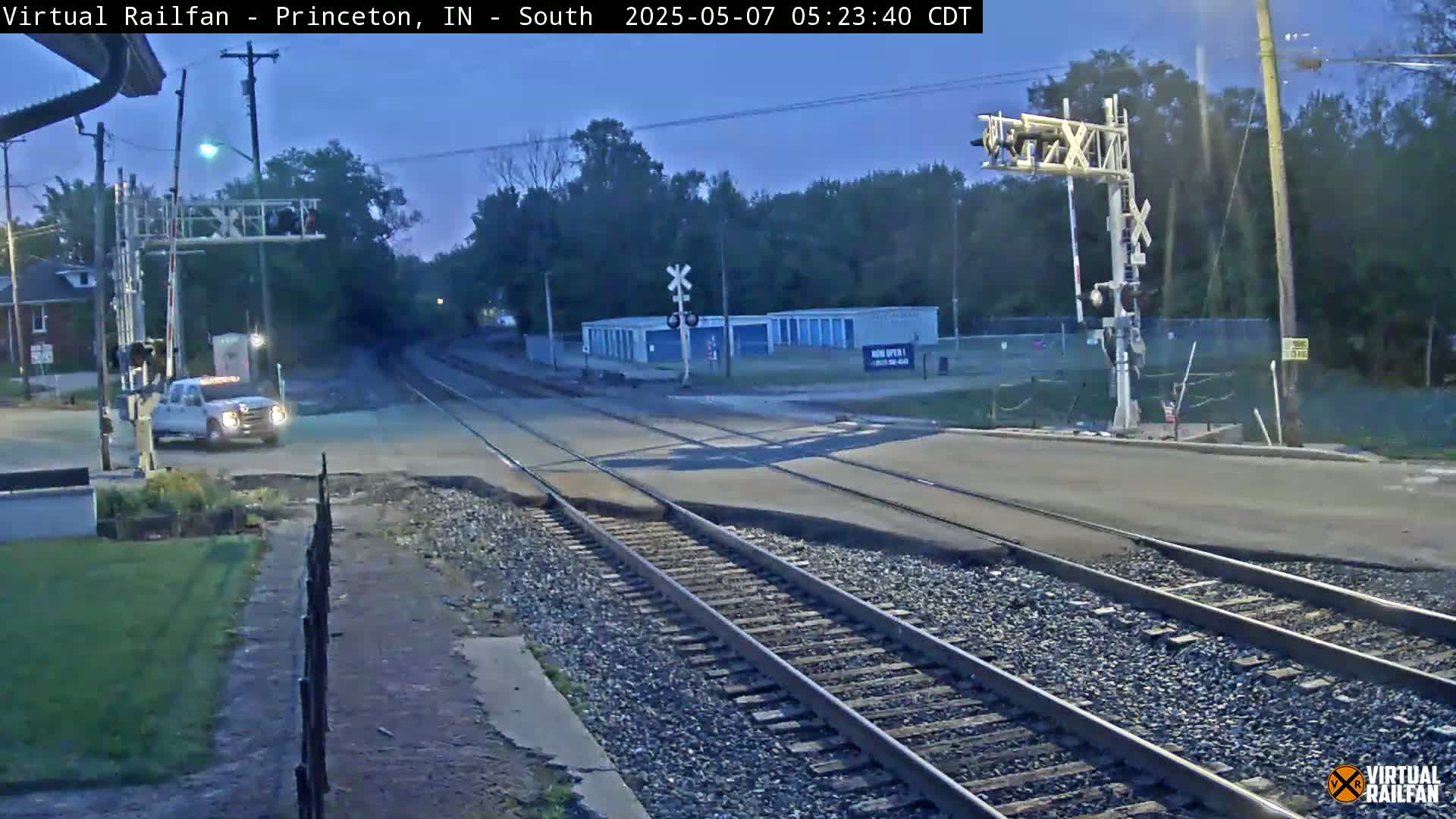 A white pickup truck sits at a railroad crossing under a twilight sky, with railroad tracks extending into the distance.