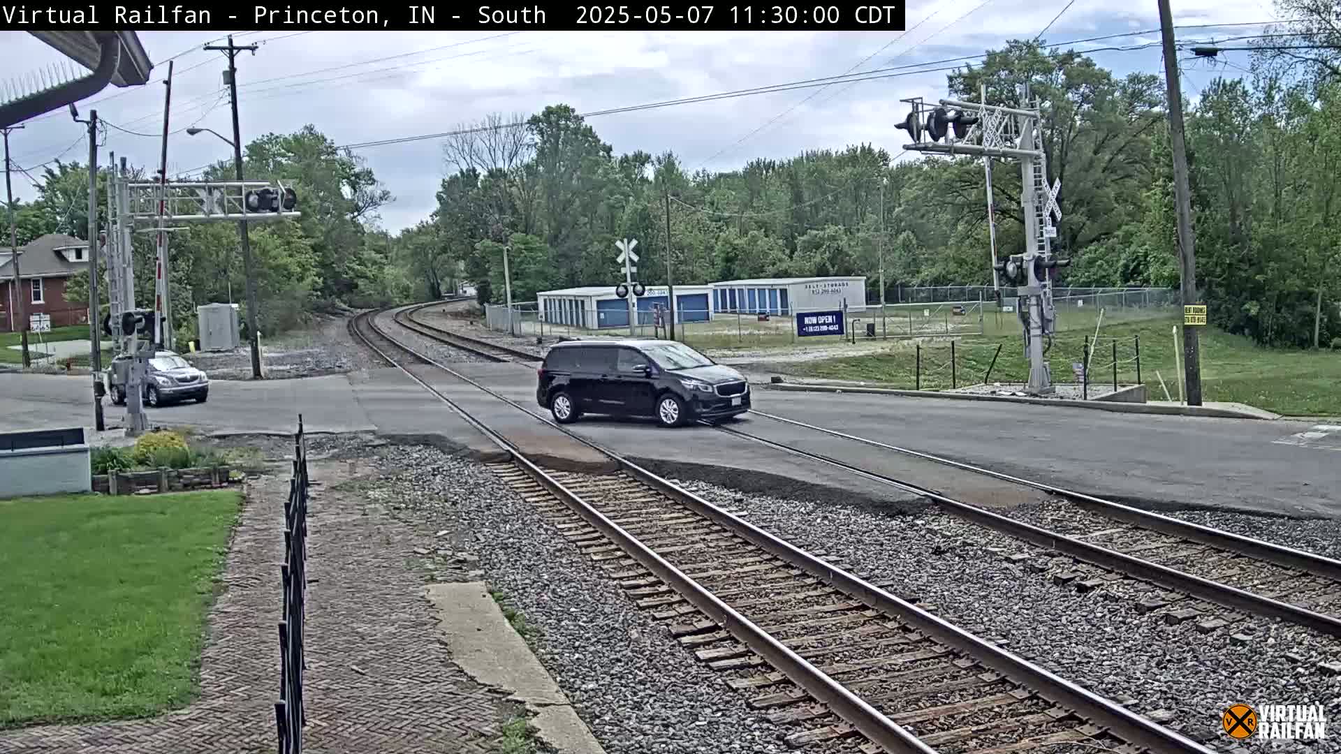 A black minivan and a silver car are crossing a railroad intersection on a partly sunny day, with trees and buildings visible in the background.