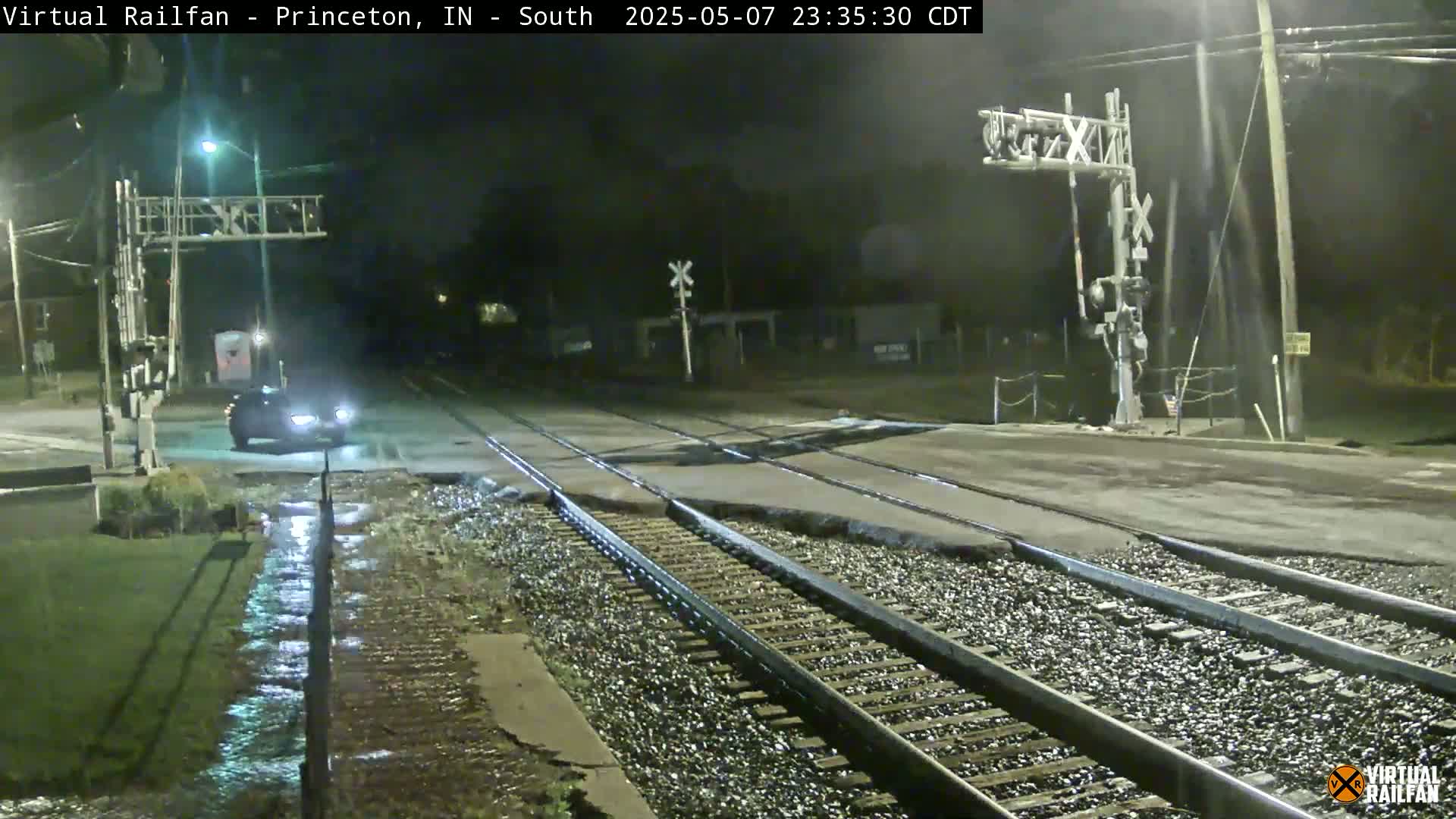 A dark-colored car drives across a wet railroad crossing at night under dim lighting.