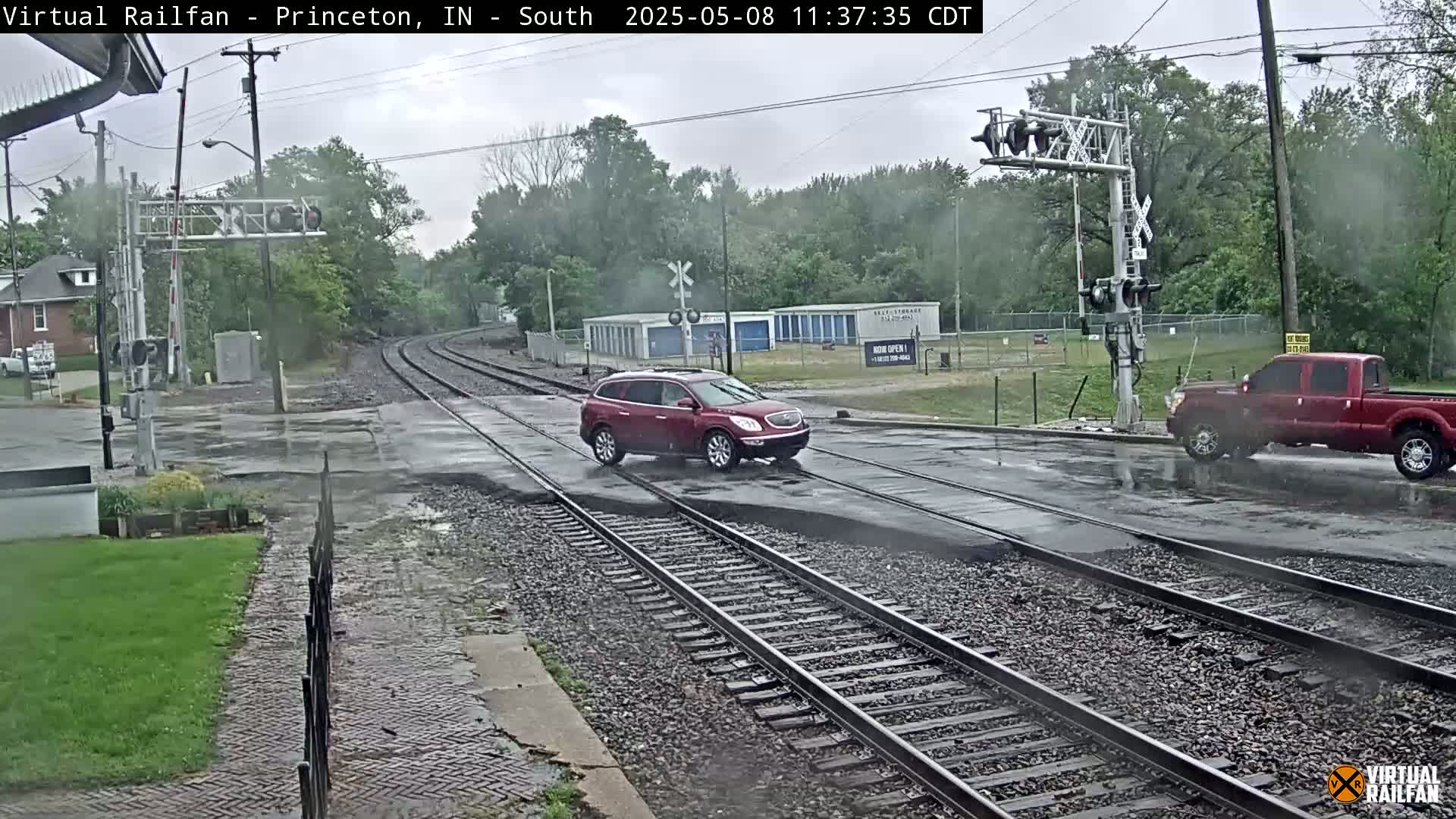 A red SUV and a red pickup truck are crossing a wet railroad crossing during a light rain.
