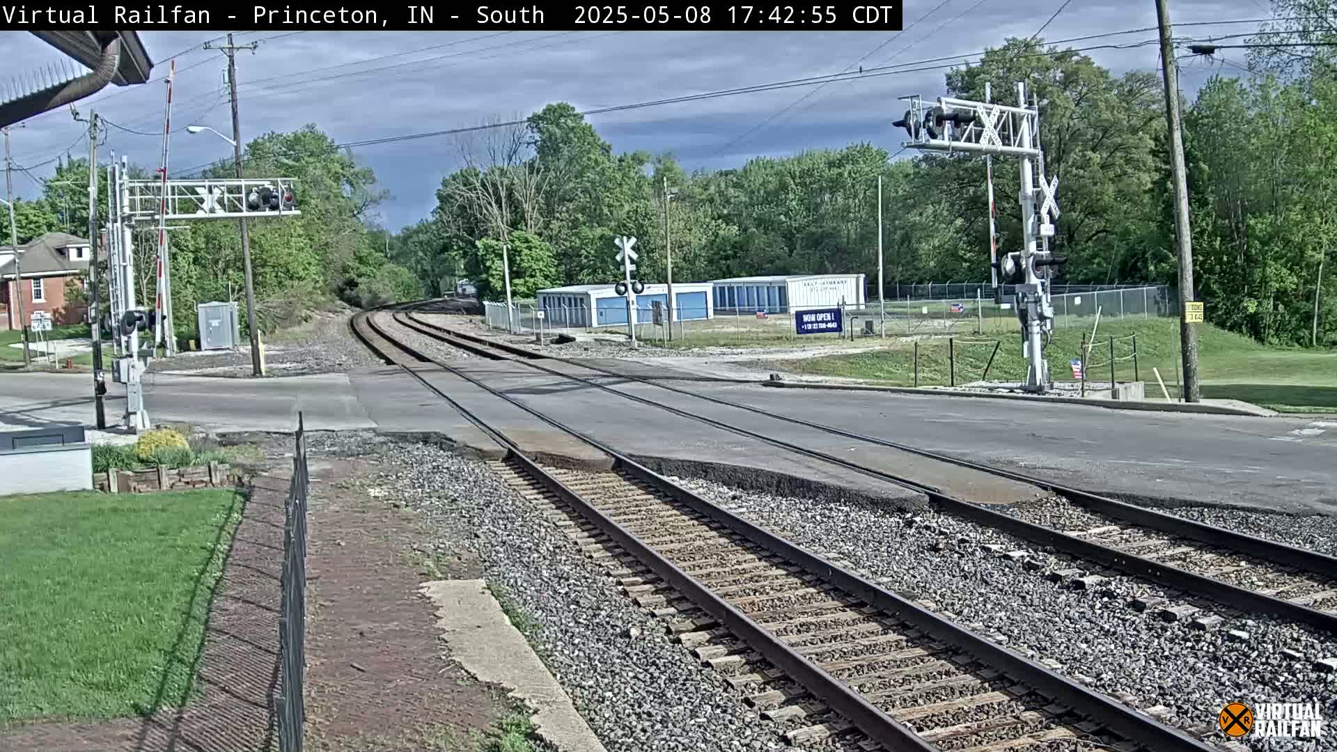 A mostly sunny day shows a railroad crossing with train tracks intersecting a road, surrounded by trees and buildings.