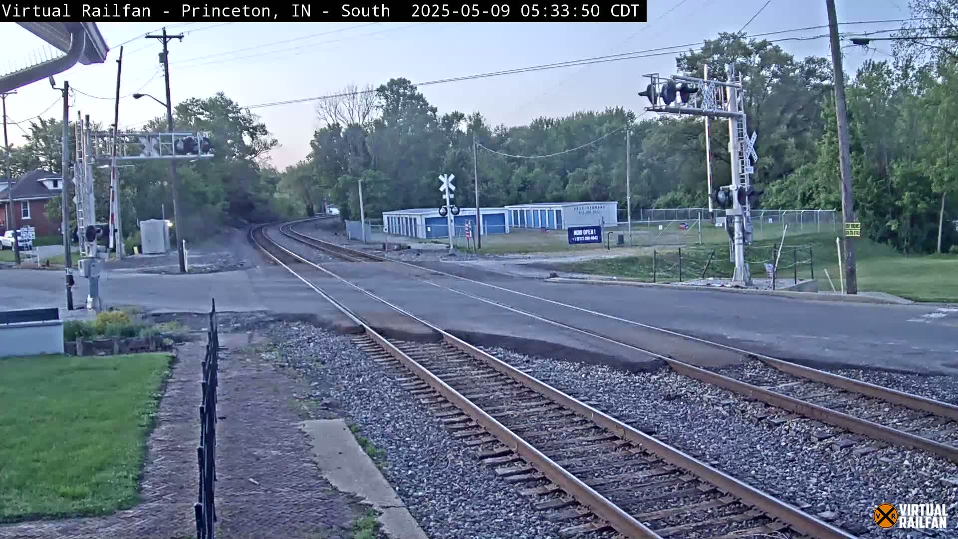 A railroad crossing with train tracks intersecting a road under a mostly clear sky, surrounded by trees and buildings.