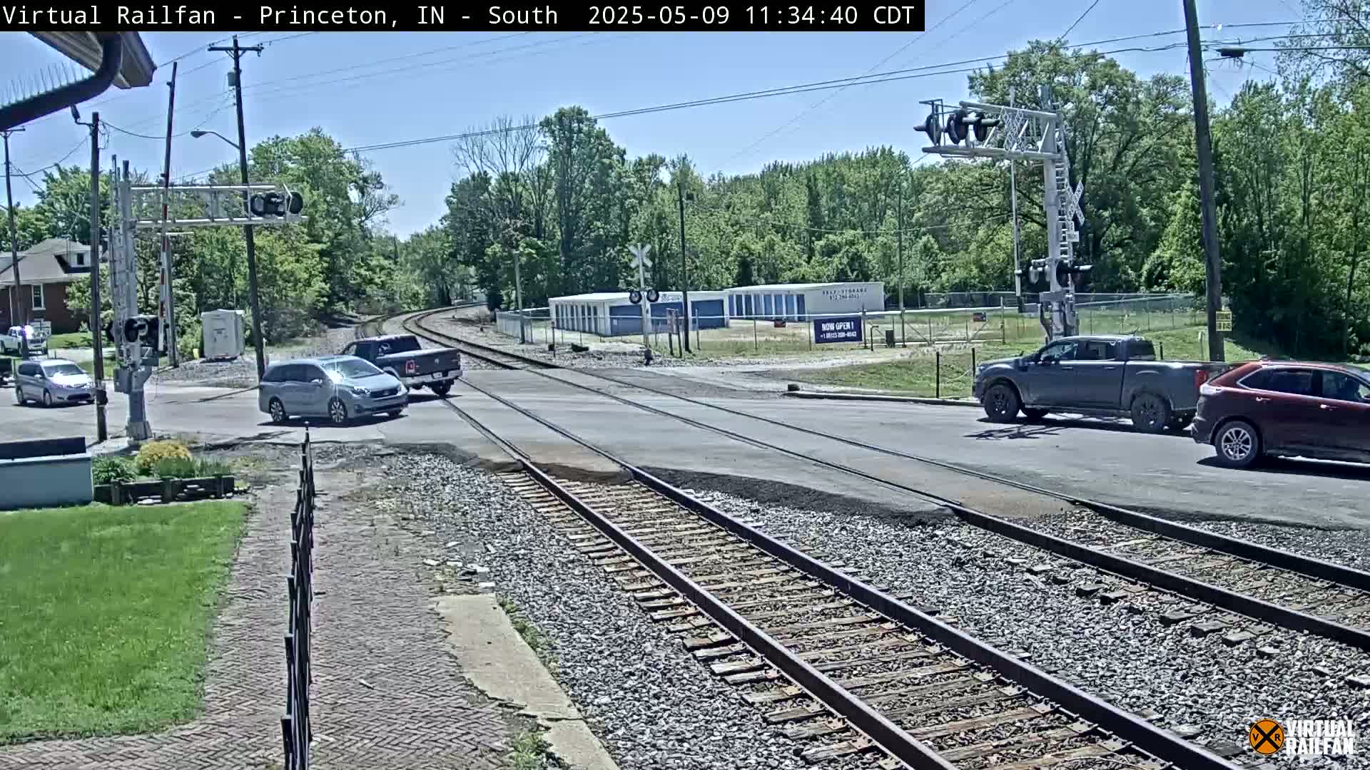 A sunny day shows several vehicles at a railroad crossing with a train track running through the middle of the intersection, surrounded by green trees and buildings.