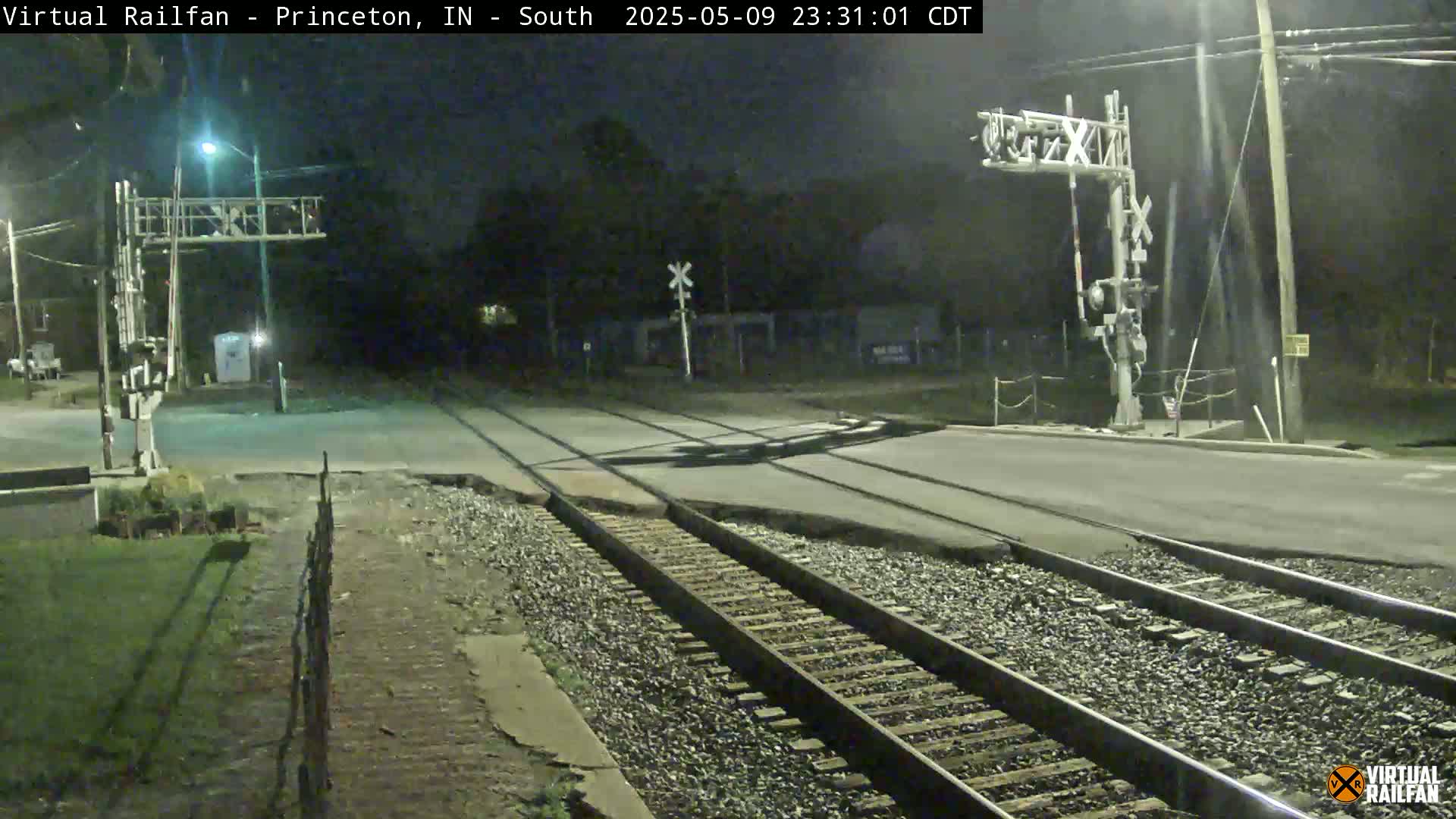 At night, railroad tracks intersect a road at a crossing equipped with signal lights, under a clear sky.