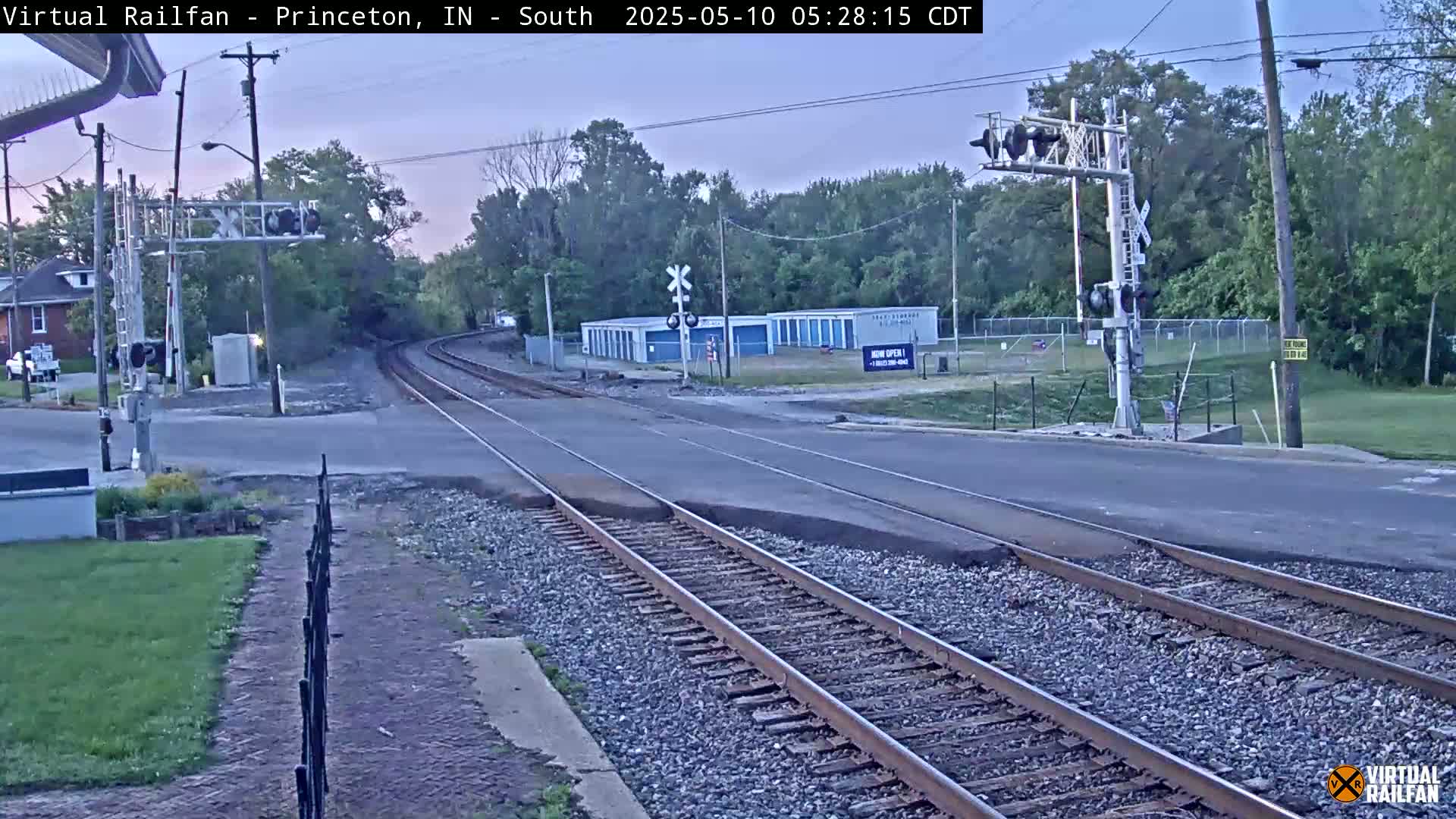 A railroad crossing with train tracks intersecting a road under a twilight sky, surrounded by trees and buildings.