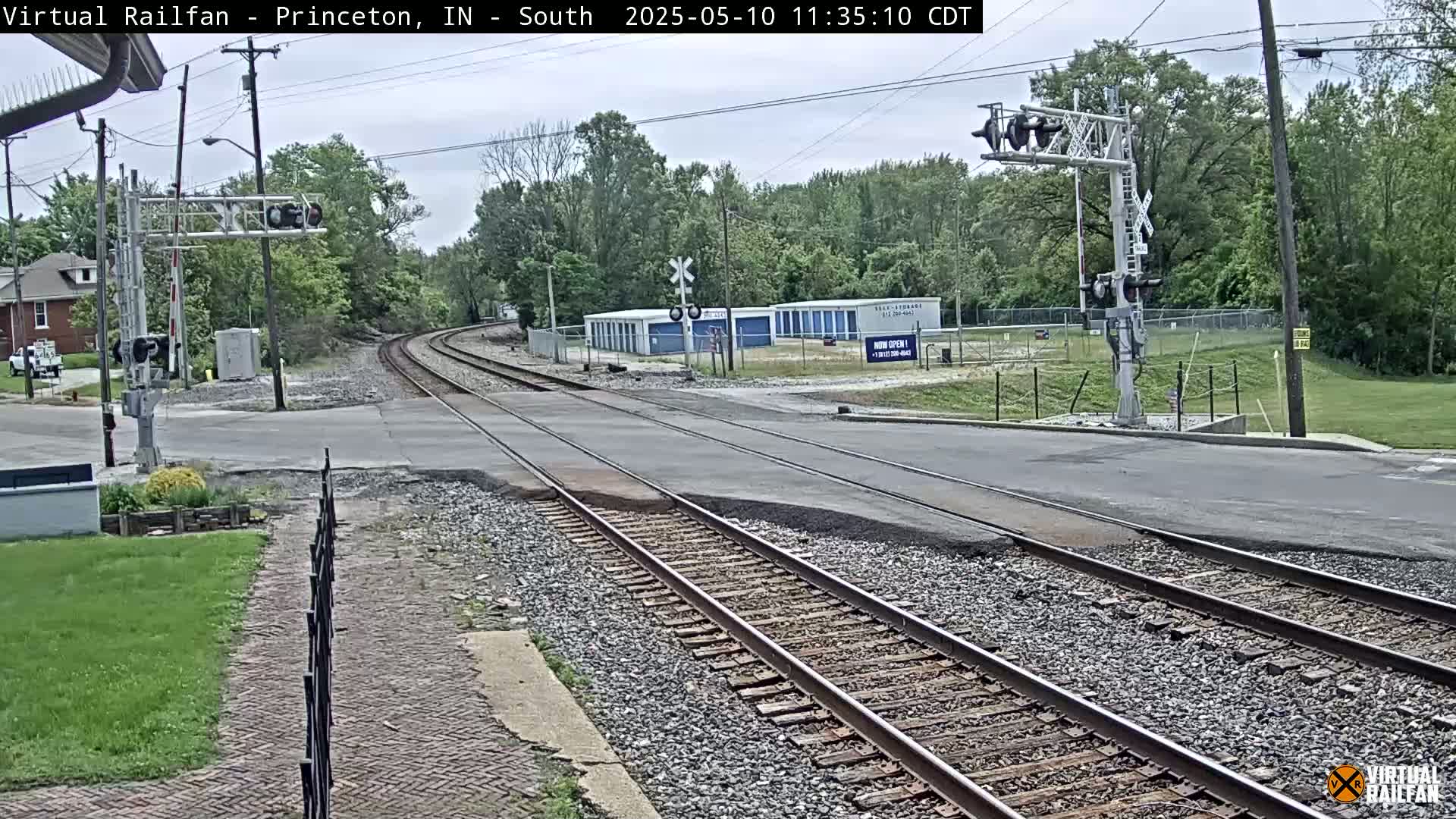 A railroad crossing with train tracks intersecting a road, under a partly cloudy sky, surrounded by greenery and a few buildings.