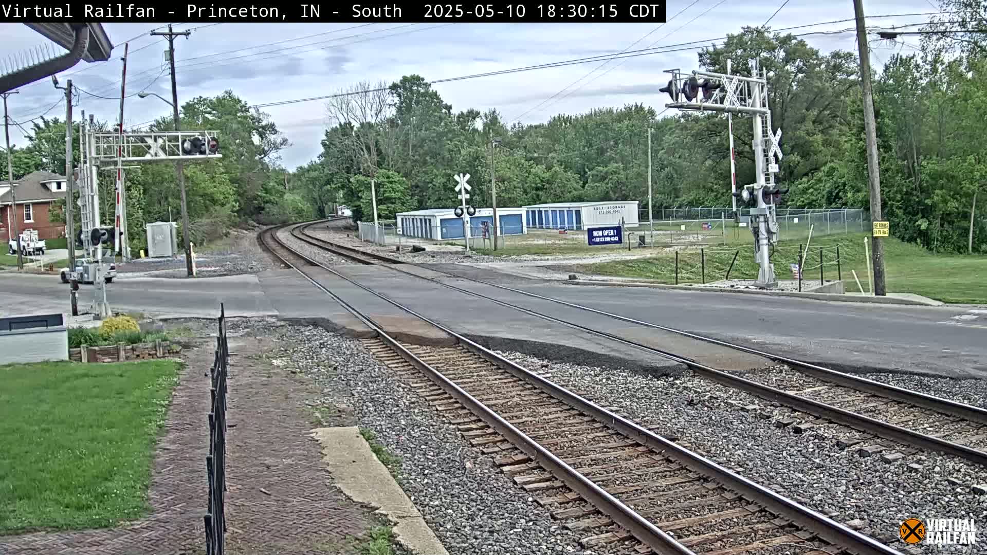 A railroad crossing with train tracks intersecting a road, under a partly cloudy sky, is surrounded by trees and a storage facility.