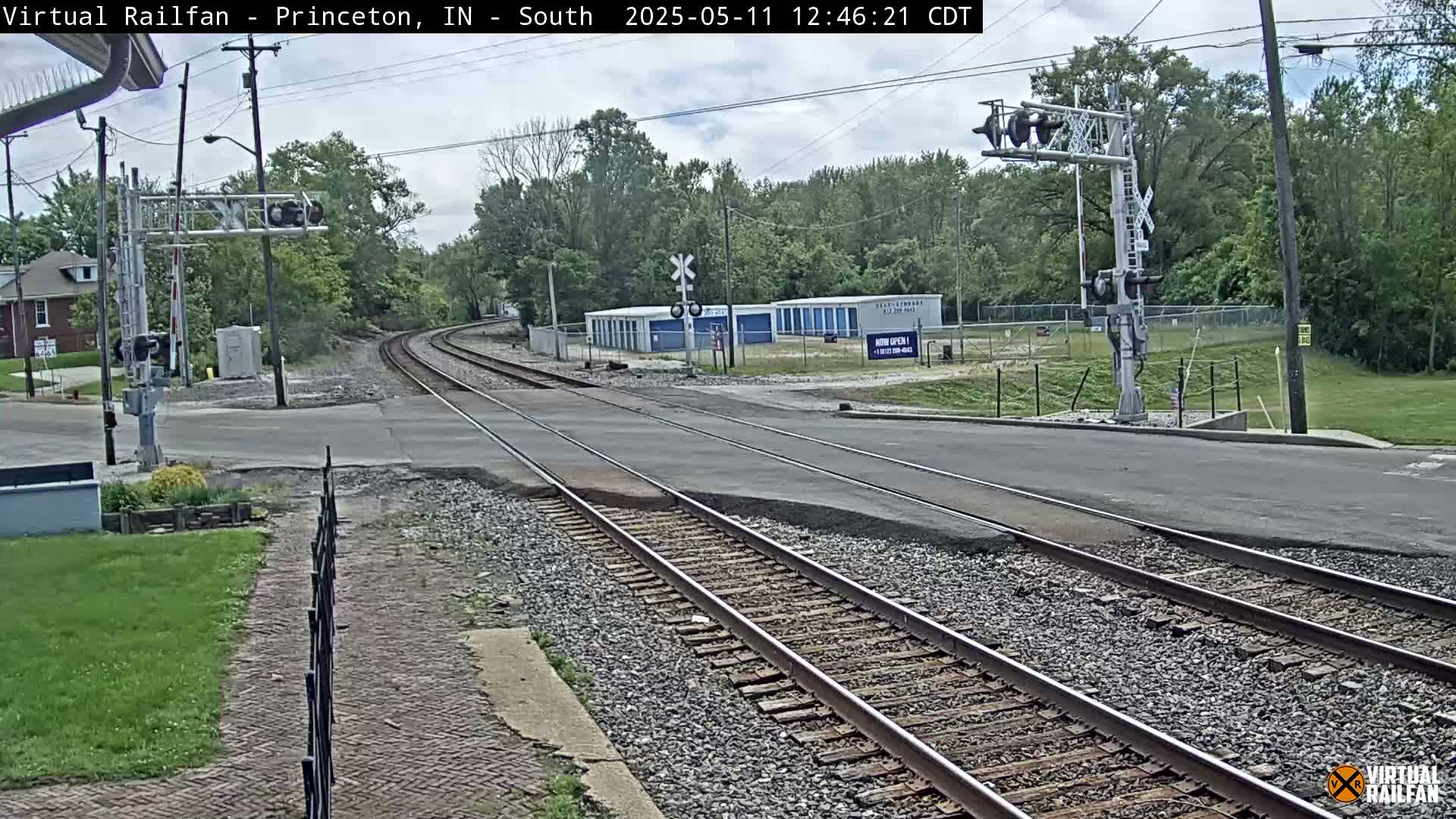 A railroad crossing with train tracks intersecting a road, under a partly cloudy sky, surrounded by trees and a storage facility.