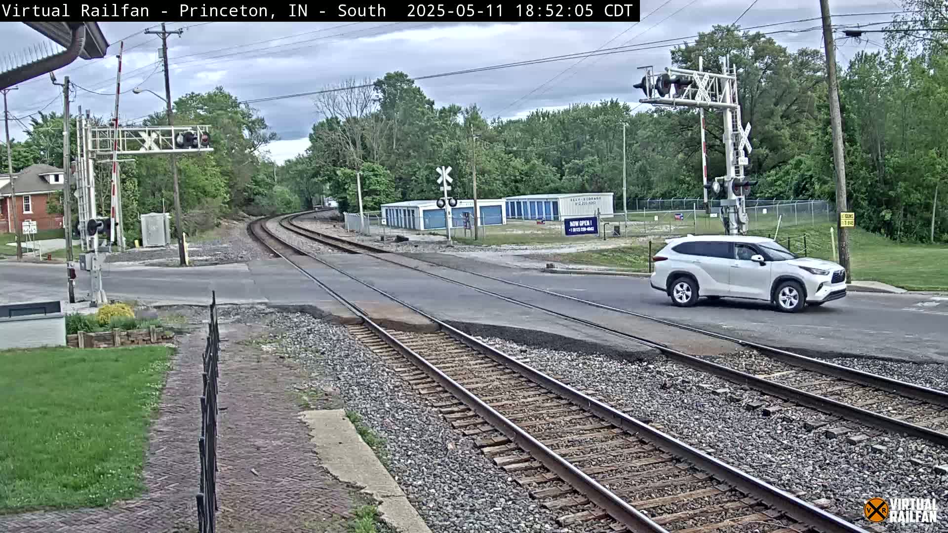 A white SUV drives across a railroad crossing under partly cloudy skies, with train tracks extending into a background of trees and storage units.