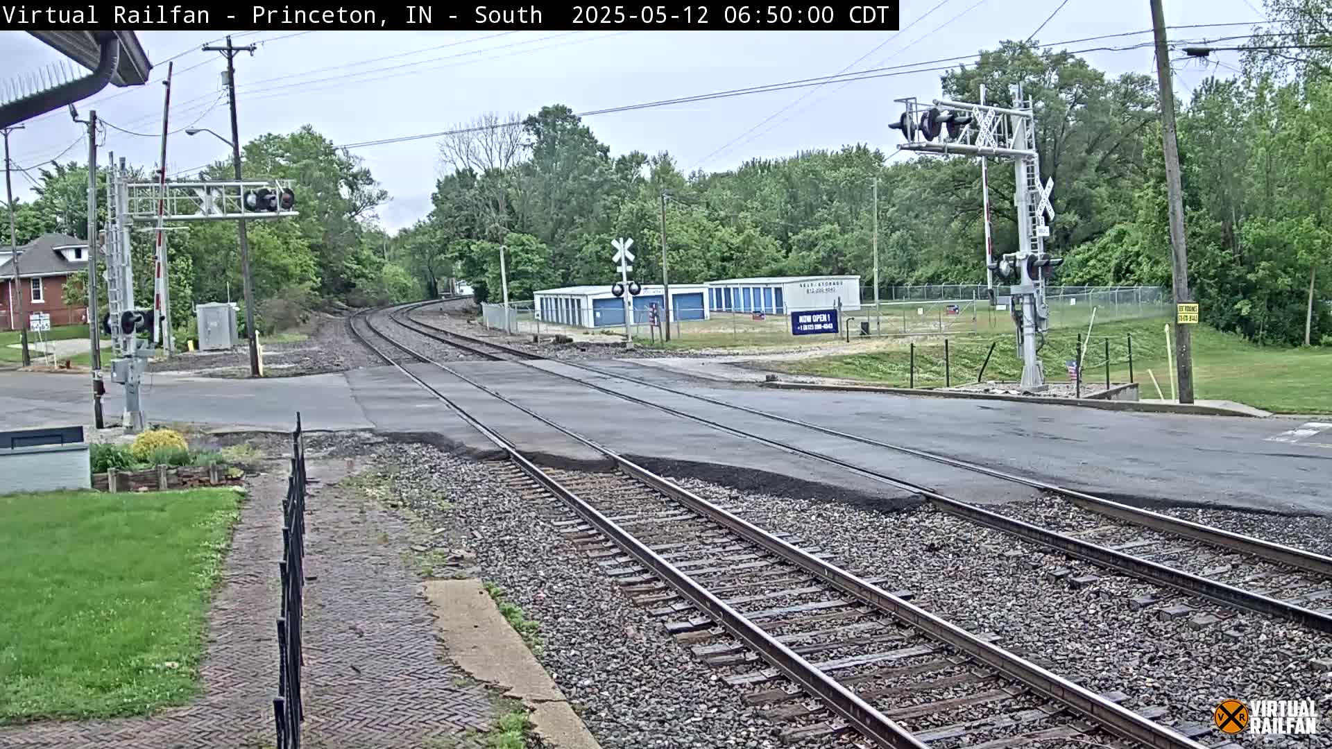 A railroad crossing with a street intersecting the tracks under an overcast sky, surrounded by green trees and grass, showing signs of recent roadwork.
