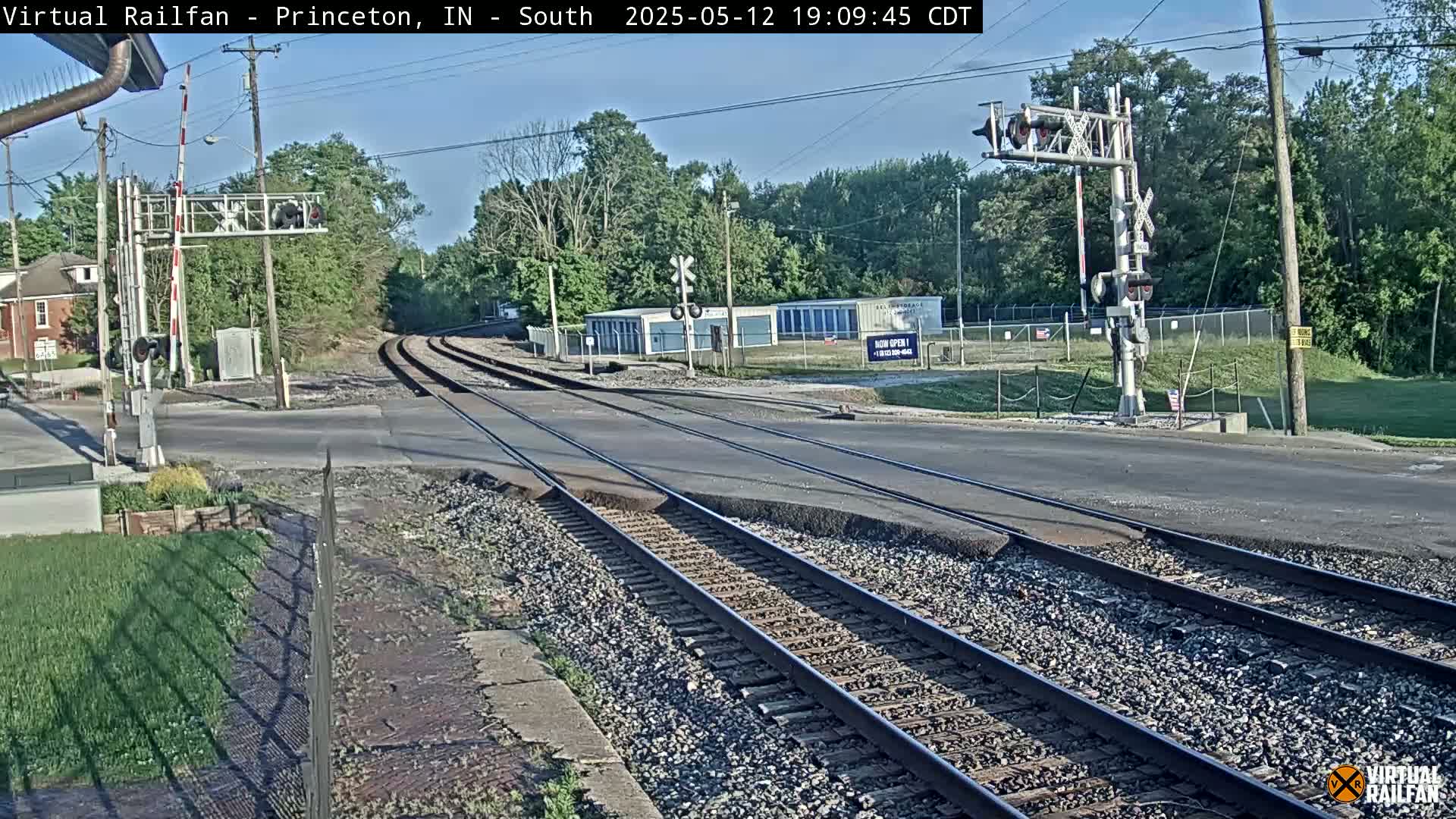A railroad crossing intersects a road under a partly sunny sky, with trees lining the background.