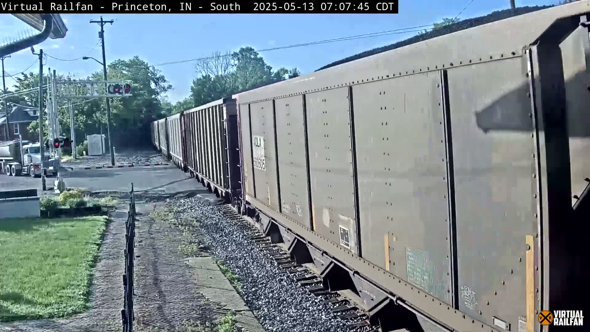 A long freight train passes a road crossing under a mostly sunny sky, with a cement truck waiting at the crossing.