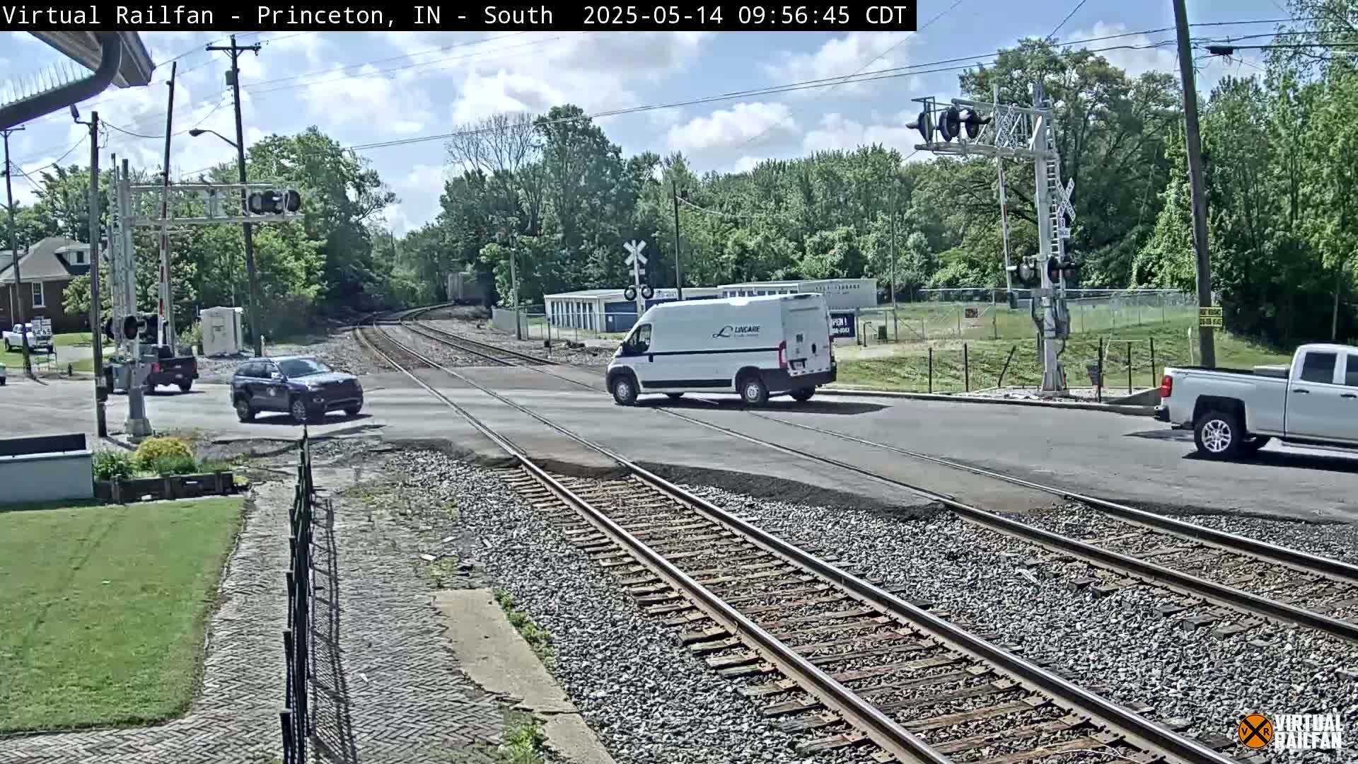 A partly sunny day shows a street intersecting train tracks, with several vehicles at the intersection near a railroad crossing signal.