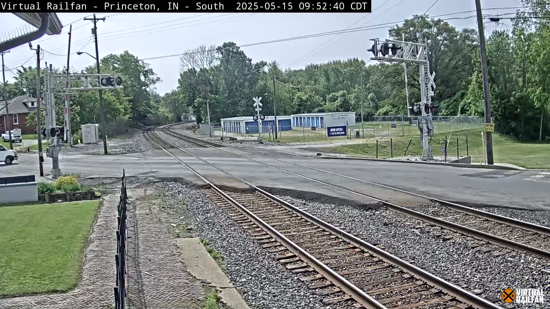 A railroad crossing intersects a road under a partly cloudy sky, with trees and storage units visible in the background.