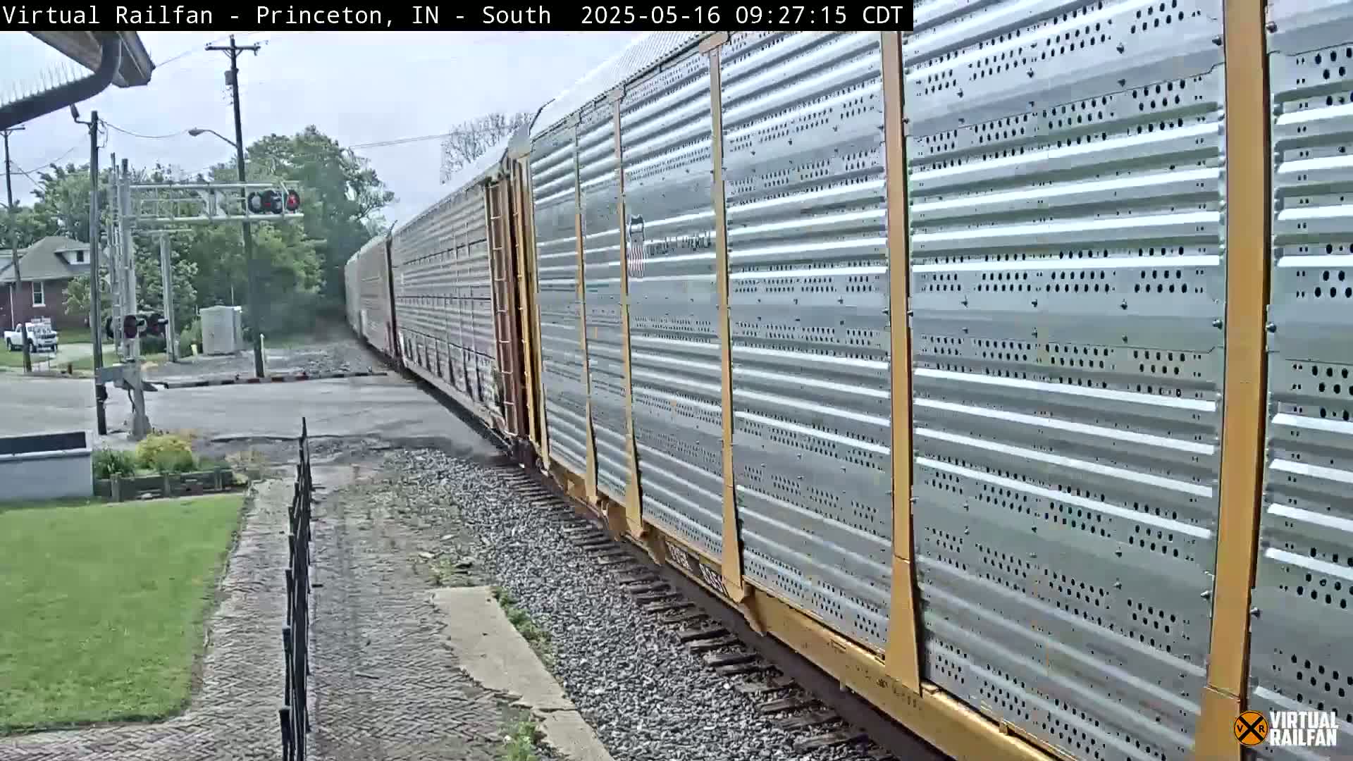 A long freight train with corrugated metal sides passes a road crossing on a partly cloudy day.