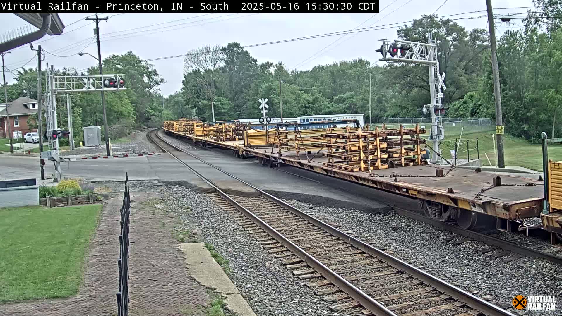 A long freight train carrying stacked yellow materials sits on a track near a road crossing under an overcast sky.